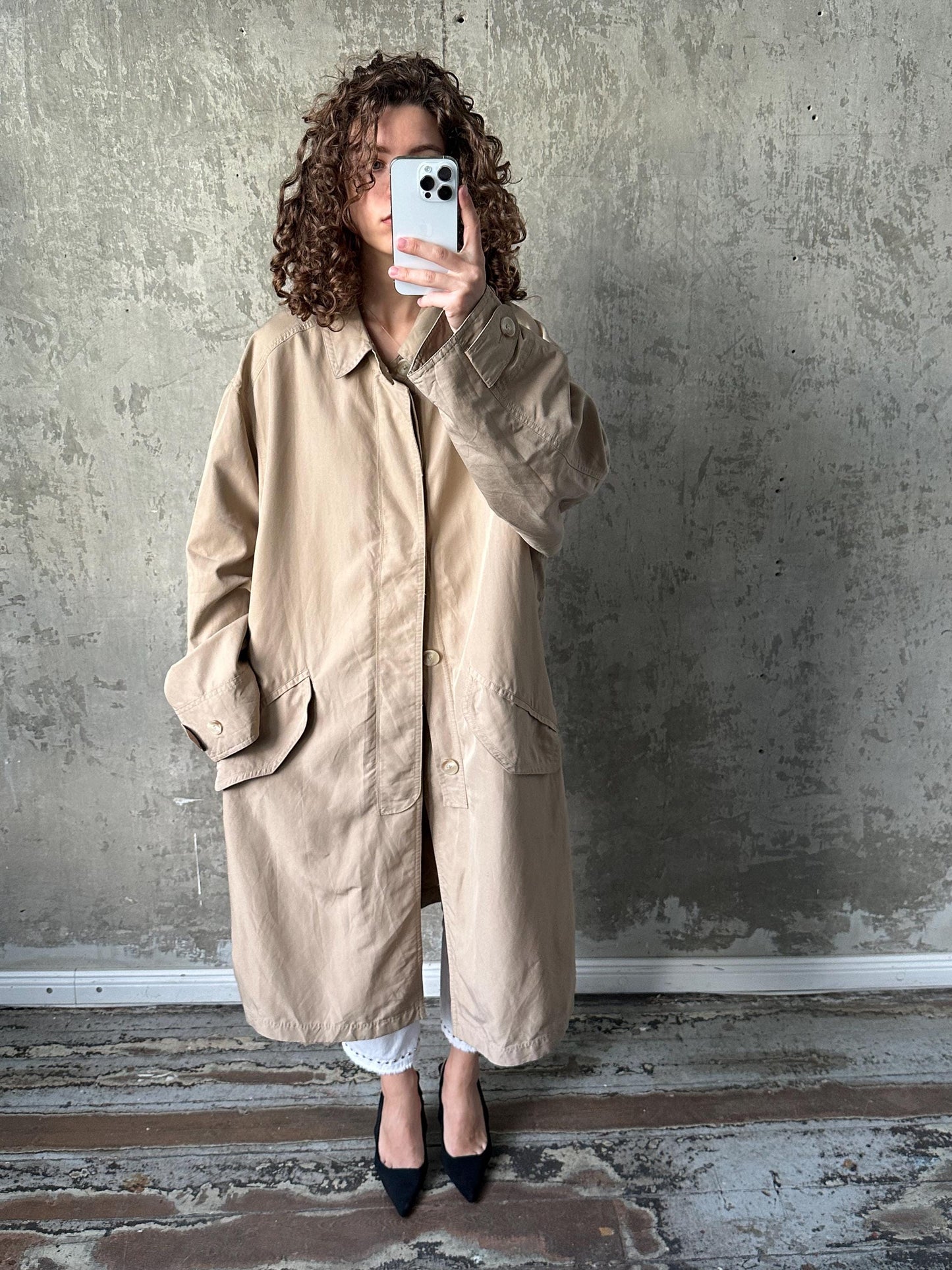 A woman with curly hair is taking a selfie with her phone, wearing a beige coat and standing in front of a concrete wall.