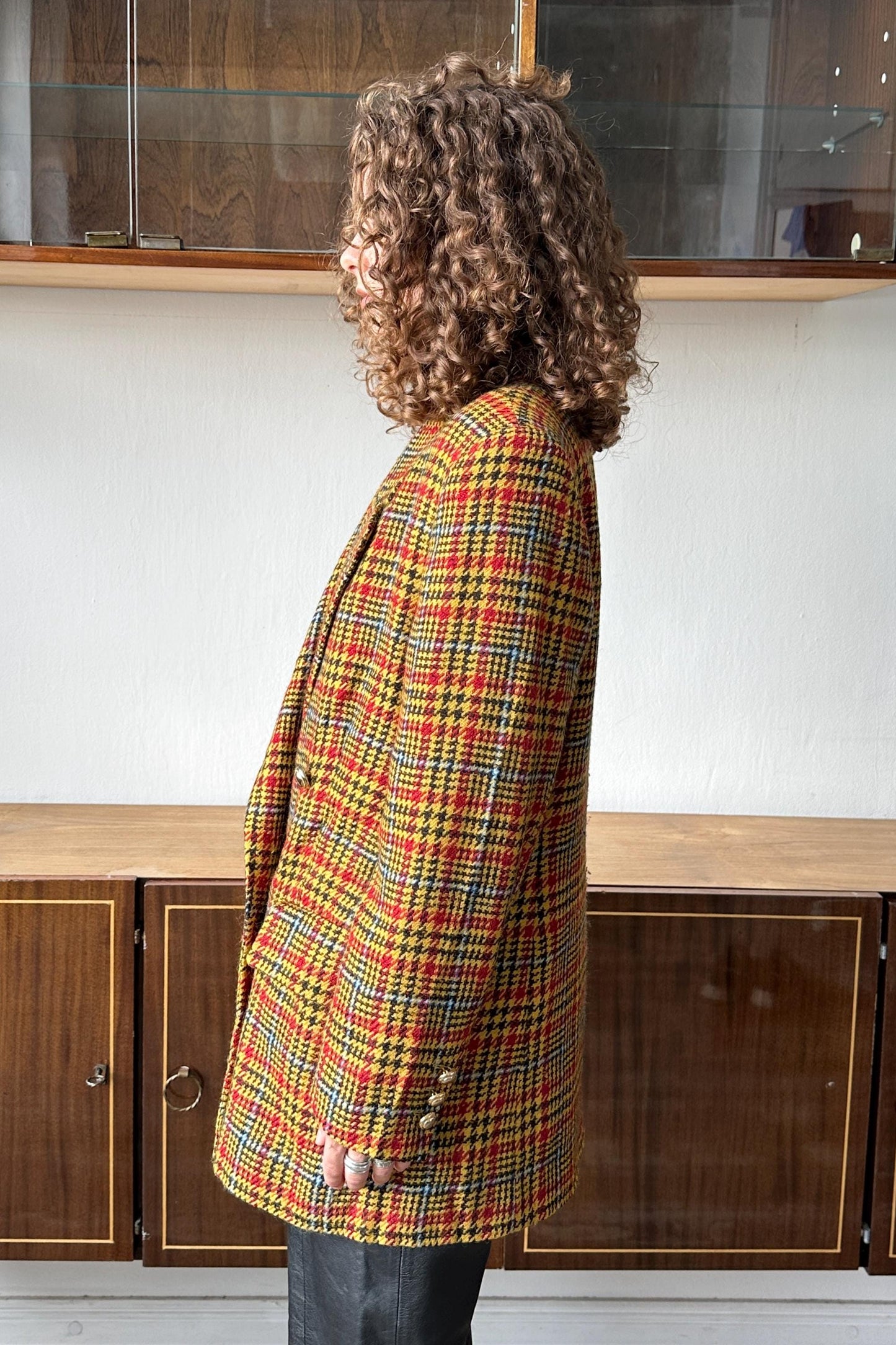 A person with curly hair wearing a colorful plaid jacket stands in front of a wooden cabinet.