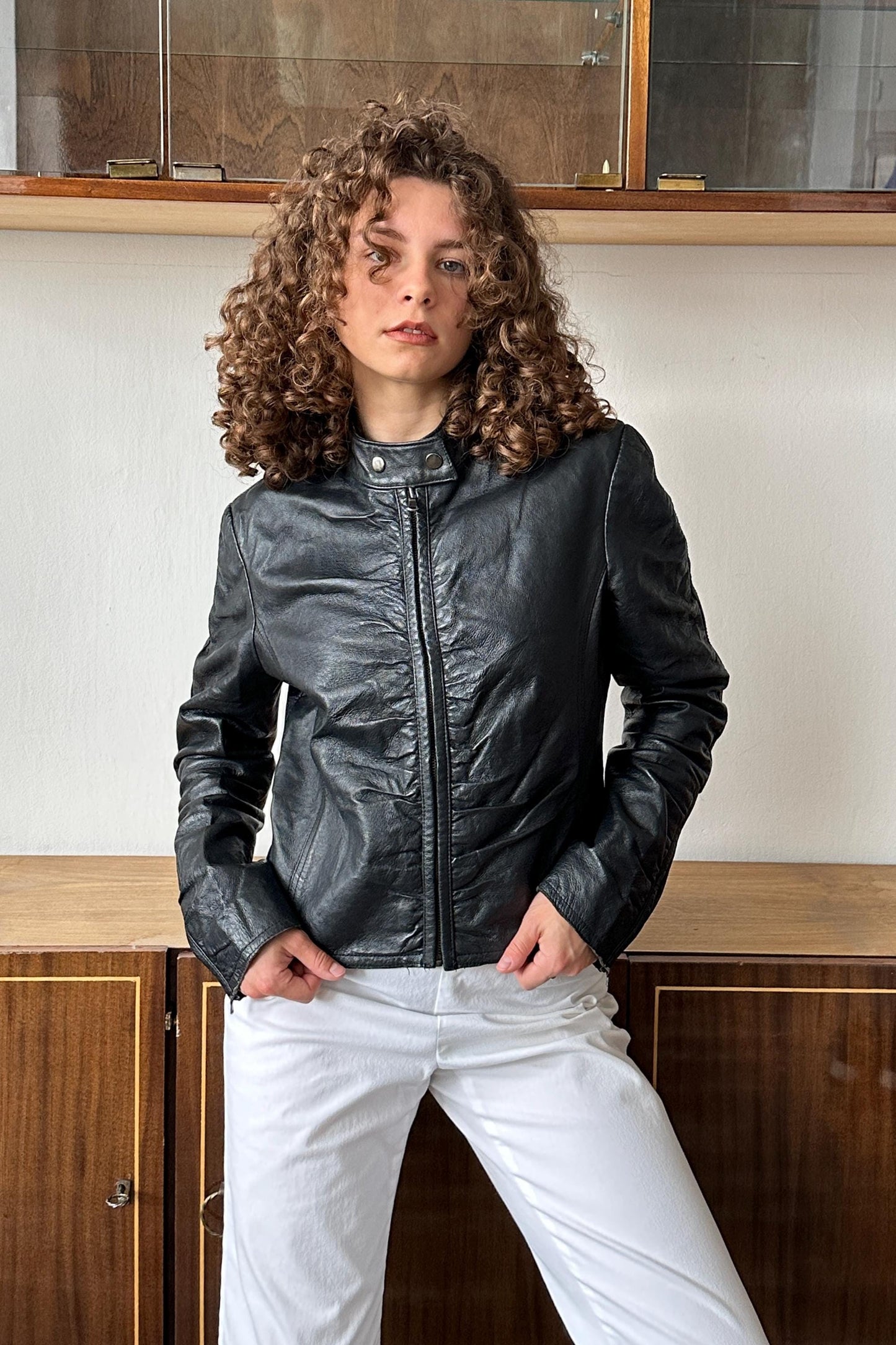 A woman with curly hair wearing a black leather jacket and white pants stands in front of a wooden cabinet.