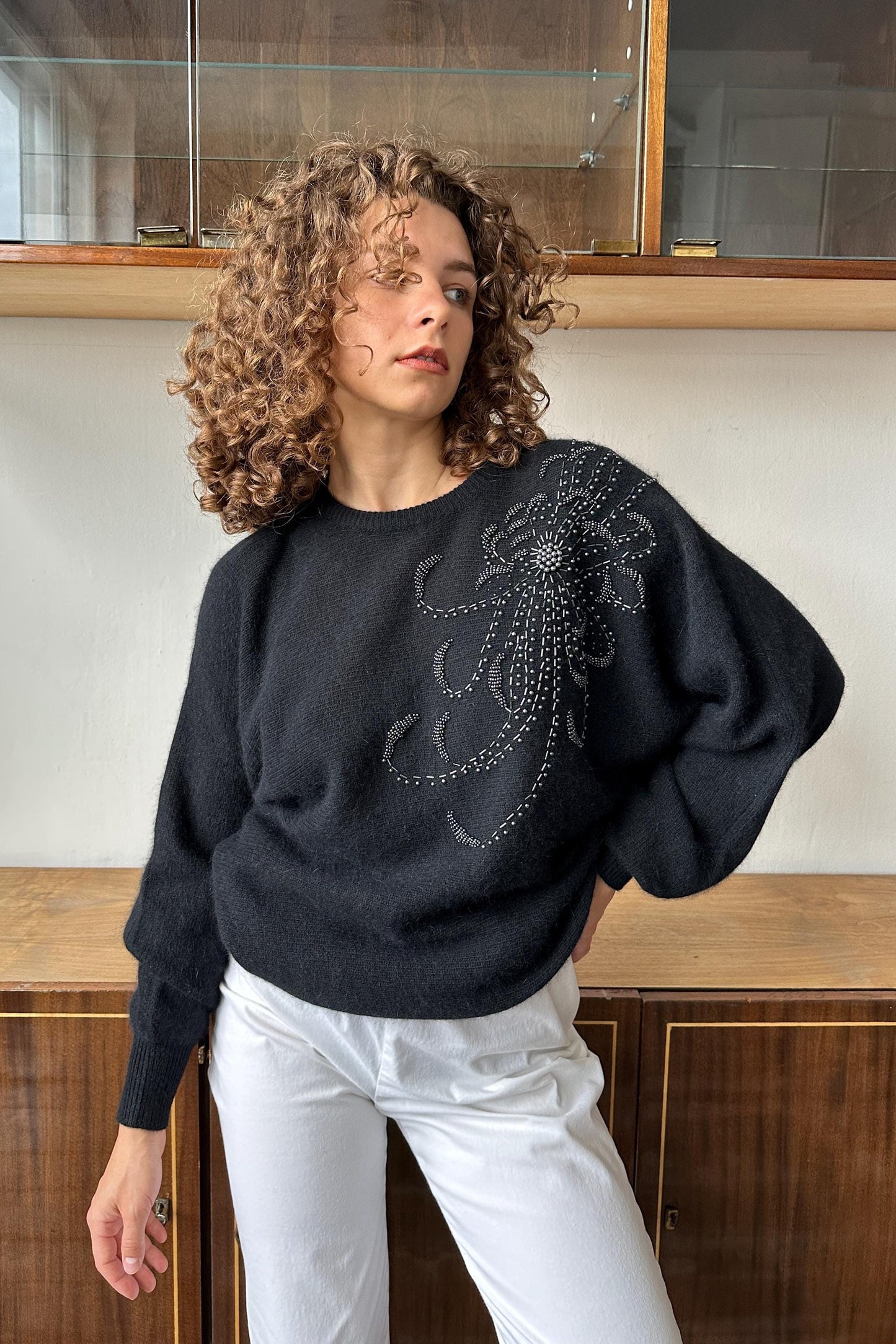 A woman with curly hair wearing a black sweater with a floral embroidered design stands in front of a wooden cabinet.