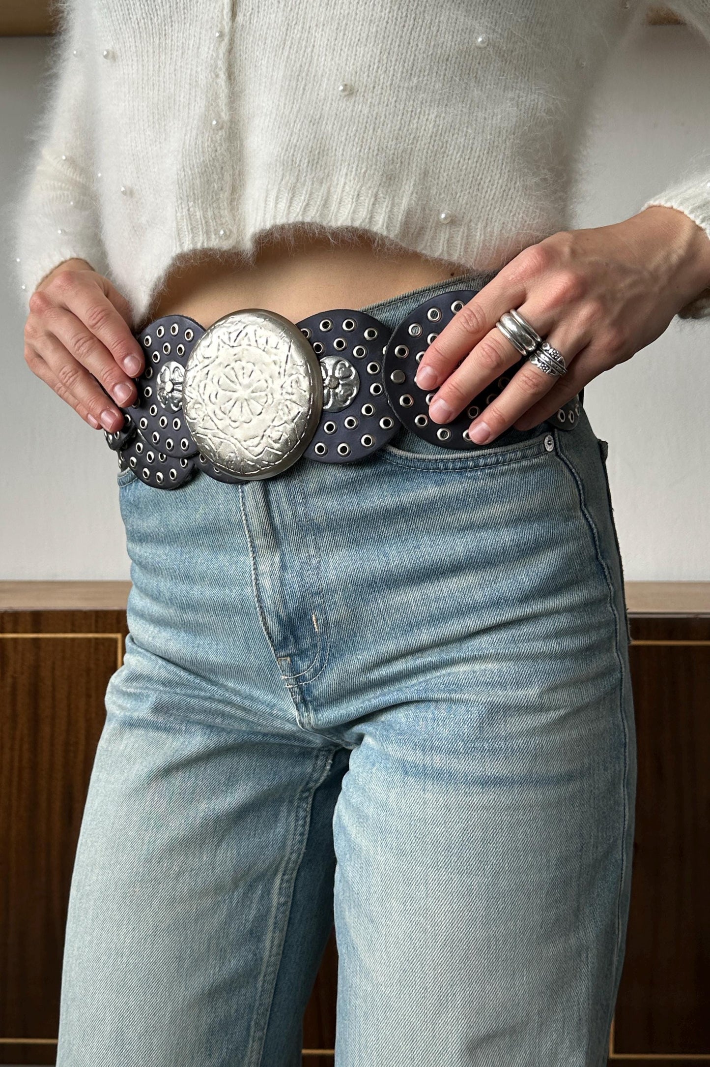 A person wearing a white sweater and blue jeans, with their hands adjusting a large, decorative belt with a metal buckle.