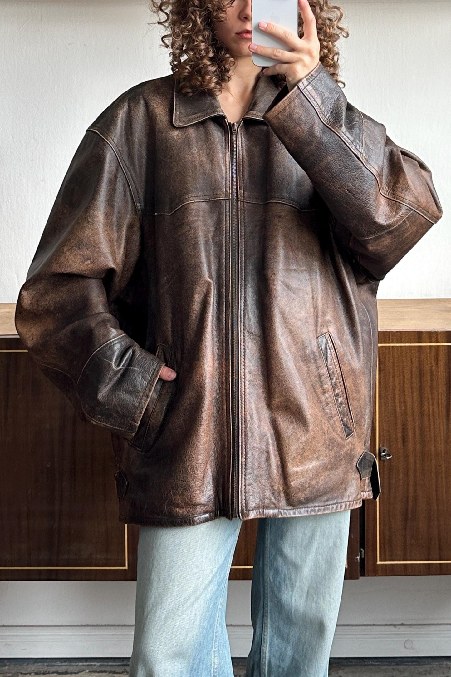 A person is taking a selfie with a brown leather jacket, standing in front of a wooden cabinet.