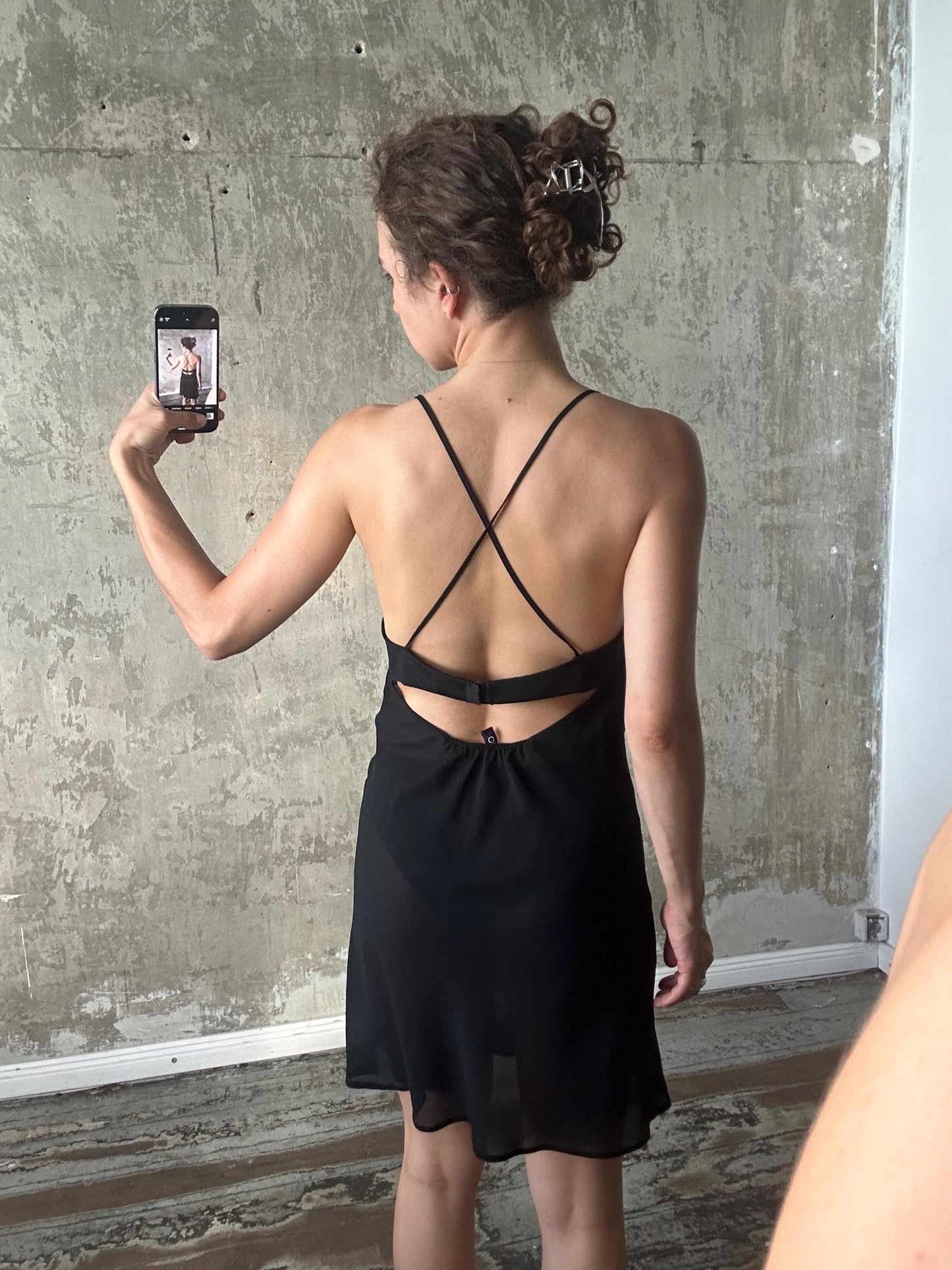 A woman in a black dress is taking a selfie with a smartphone, capturing her back view.
