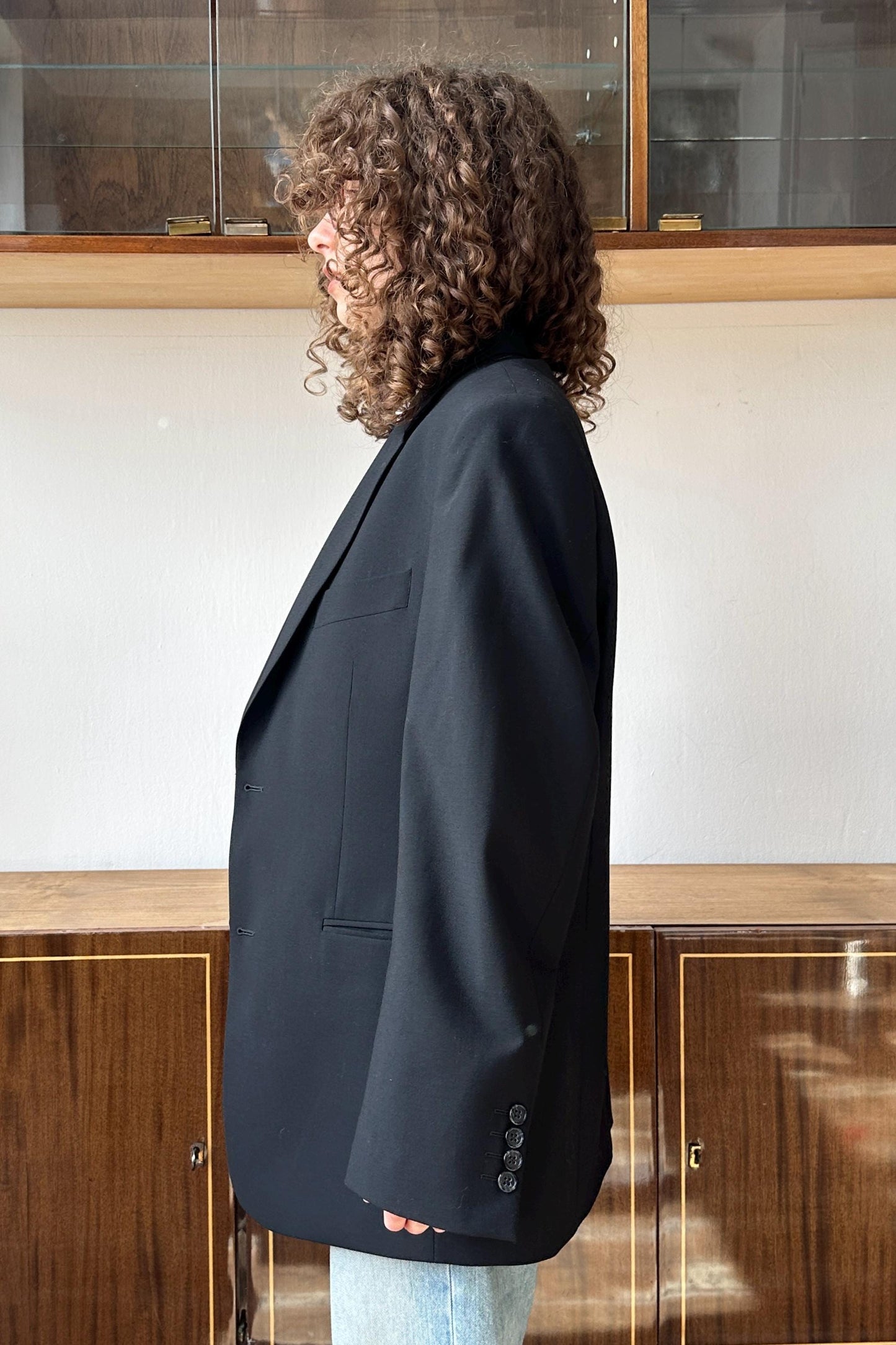 A person with curly hair wearing a black blazer stands in front of a wooden cabinet.