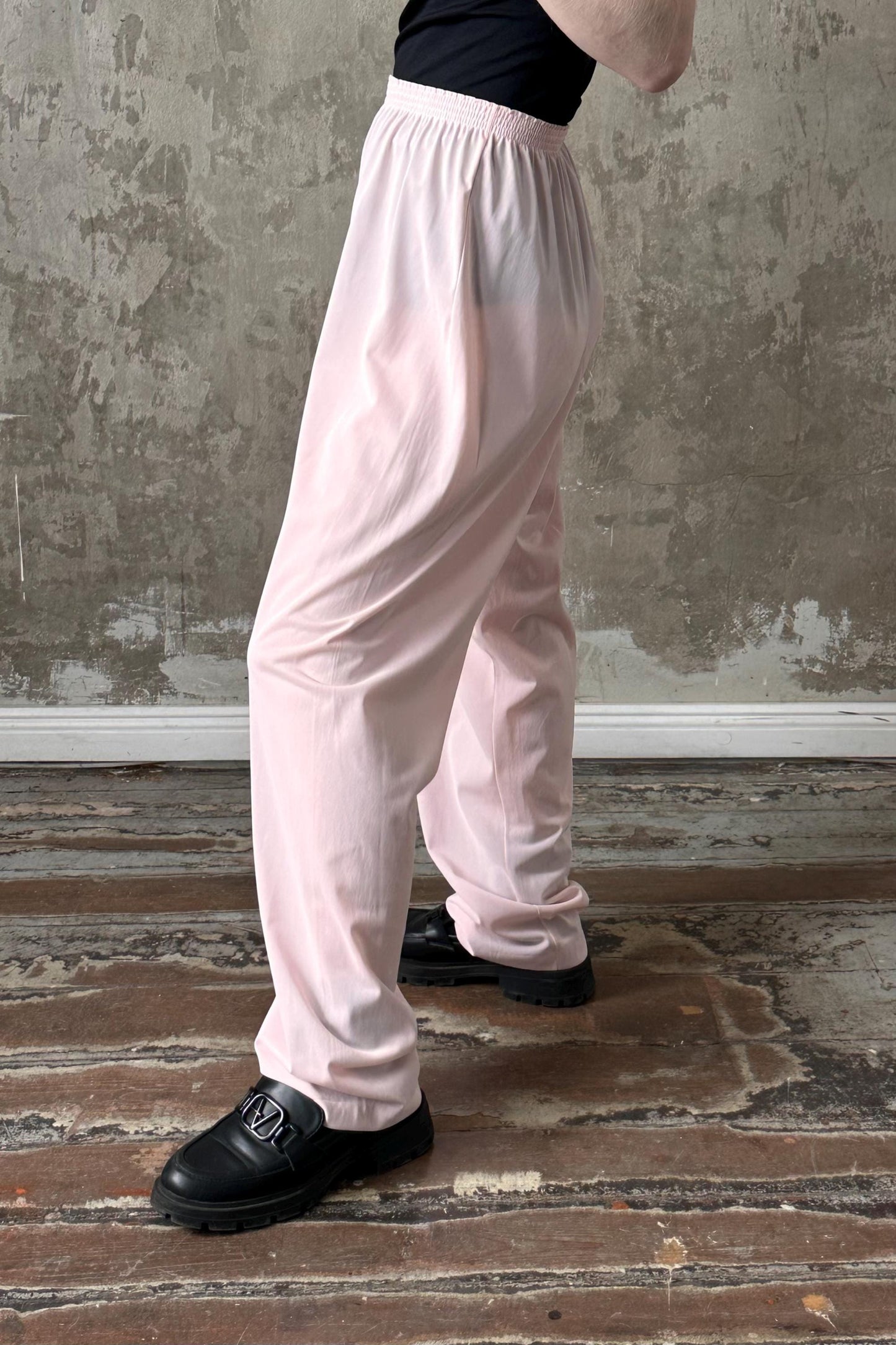 A person wearing white pants and black shoes stands in front of a concrete wall.