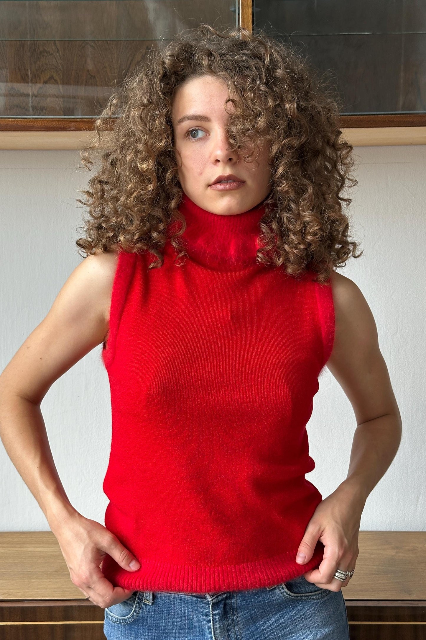 A woman with curly hair wearing a red sleeveless top and blue jeans stands in front of a wooden wall.