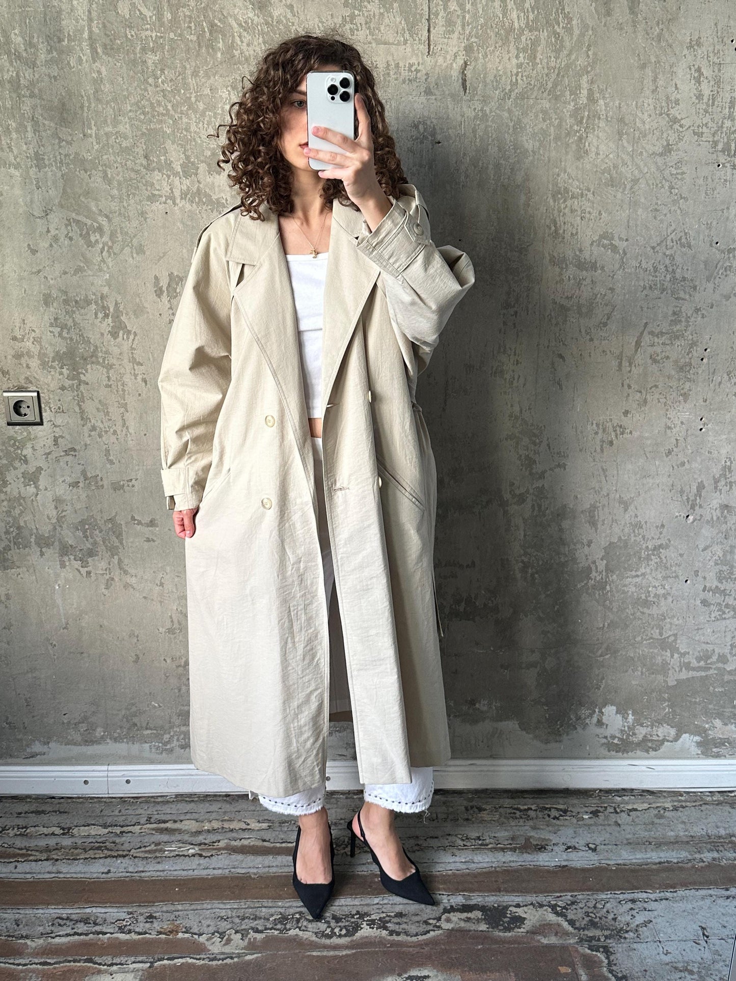 A woman in a beige trench coat is taking a selfie with a smartphone, standing against a concrete wall.