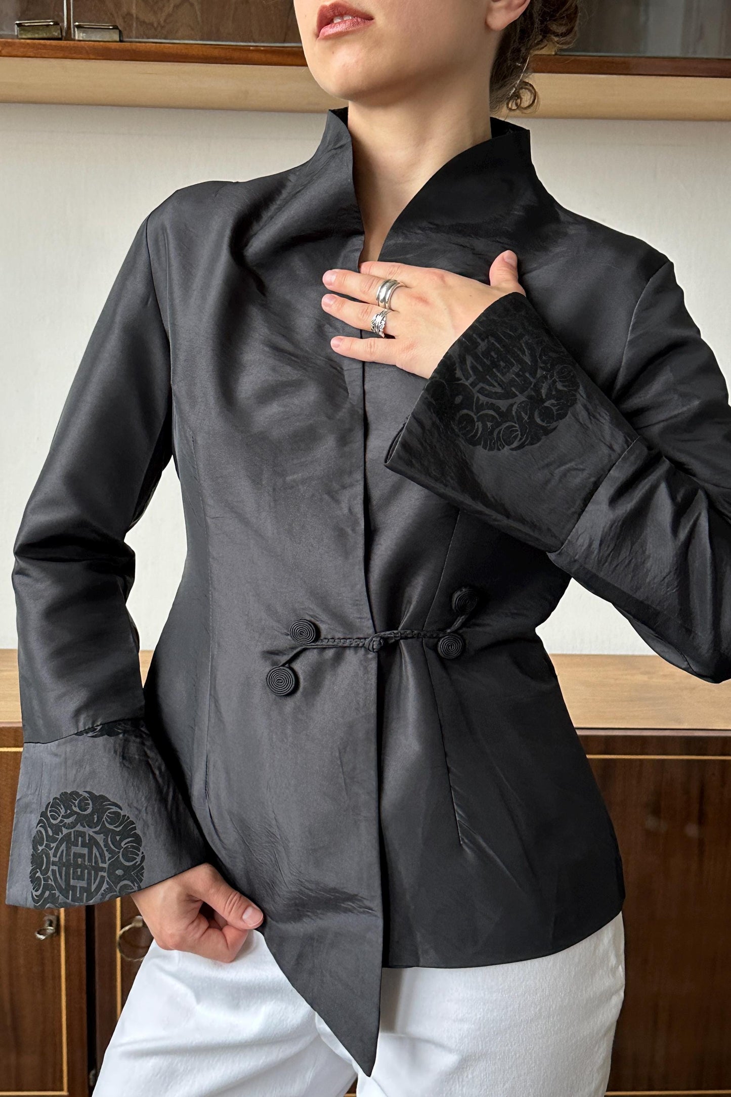 A woman wearing a black jacket with a high collar and a black floral pattern on the sleeves, posing with her hand on her chest.