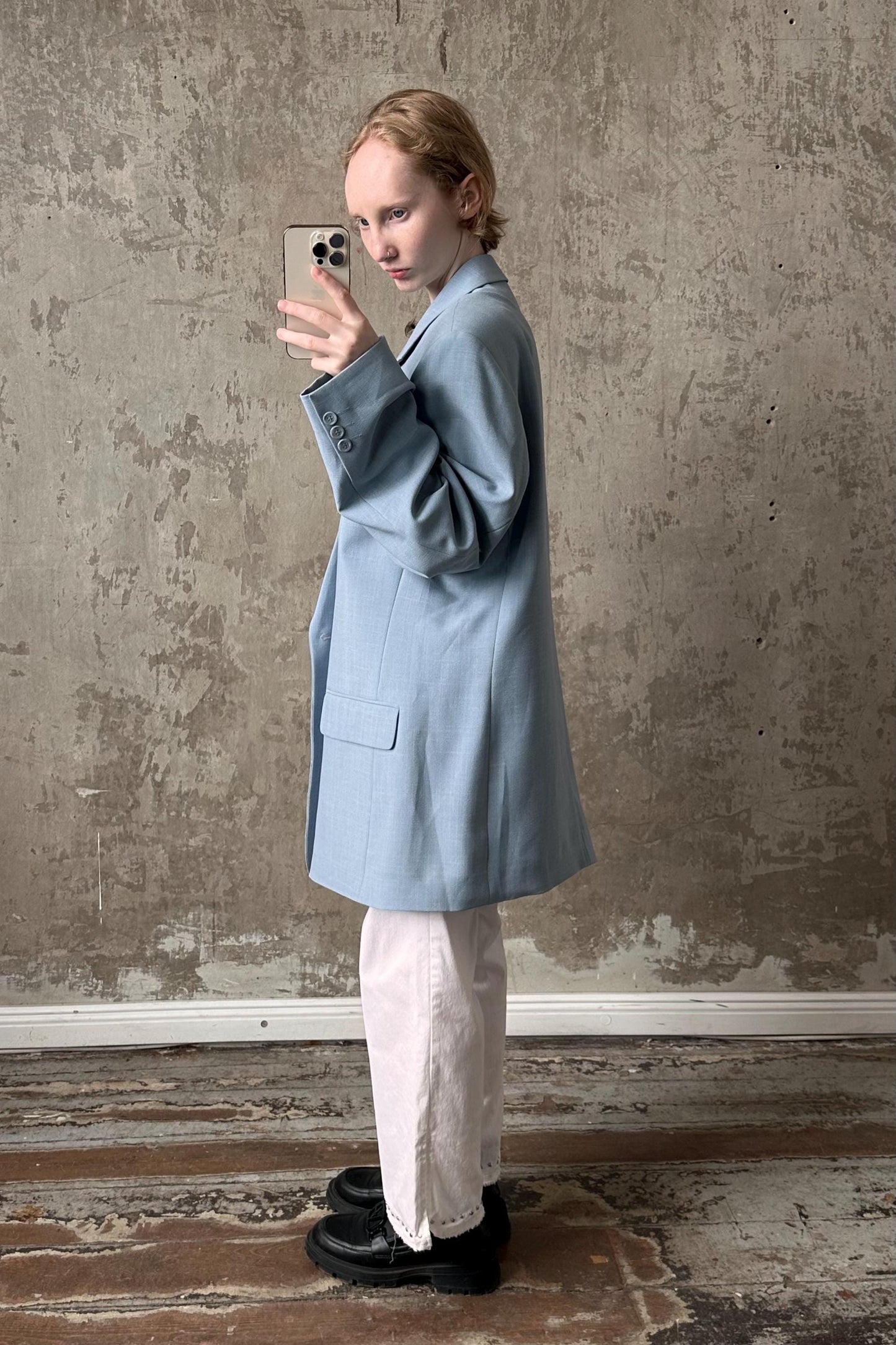 A person in a light blue blazer and white pants is taking a selfie with a smartphone, standing in front of a concrete wall.