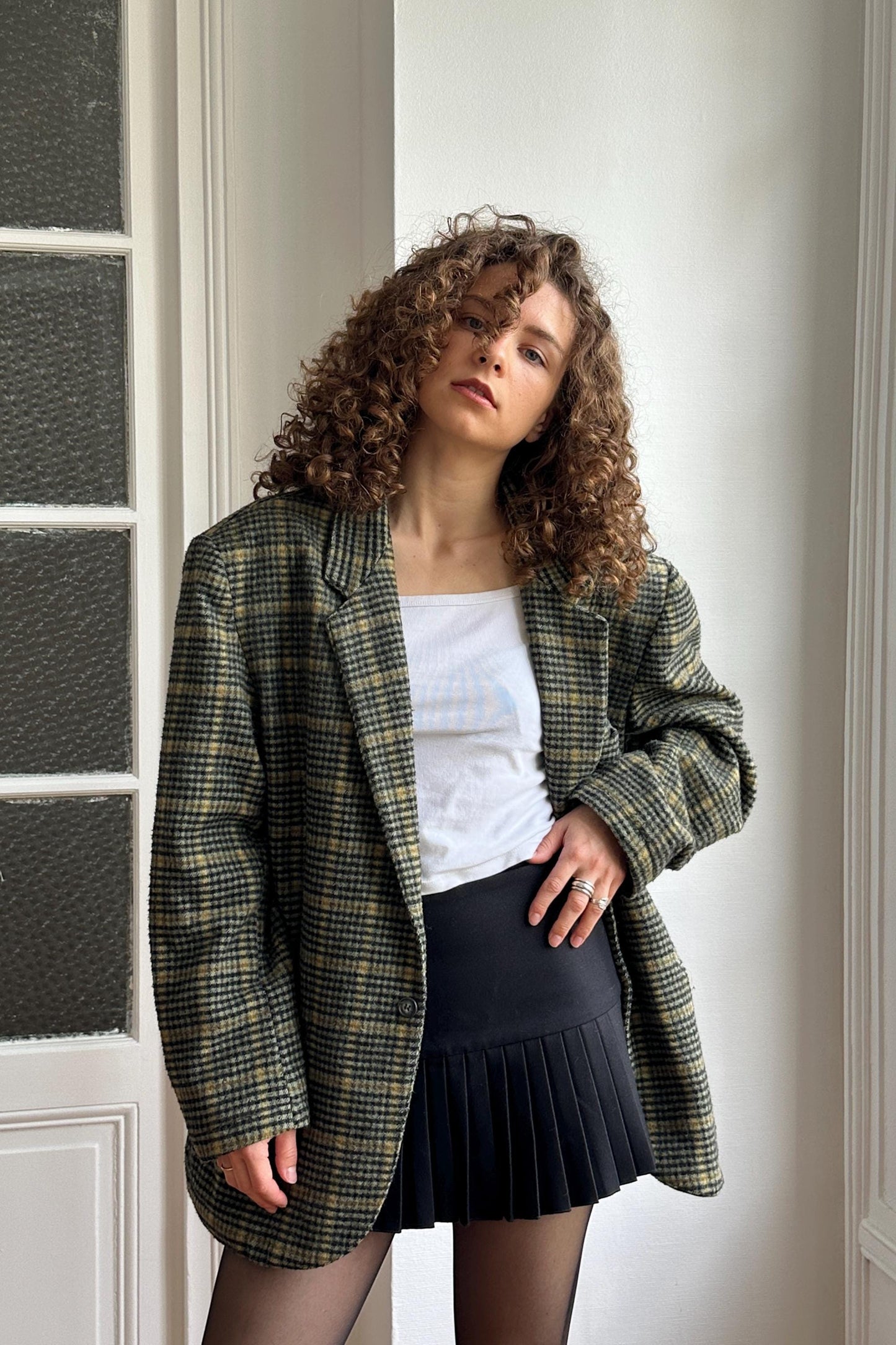 A young woman with curly hair stands in front of a white wall, wearing a plaid jacket, a white top, and a black pleated skirt.