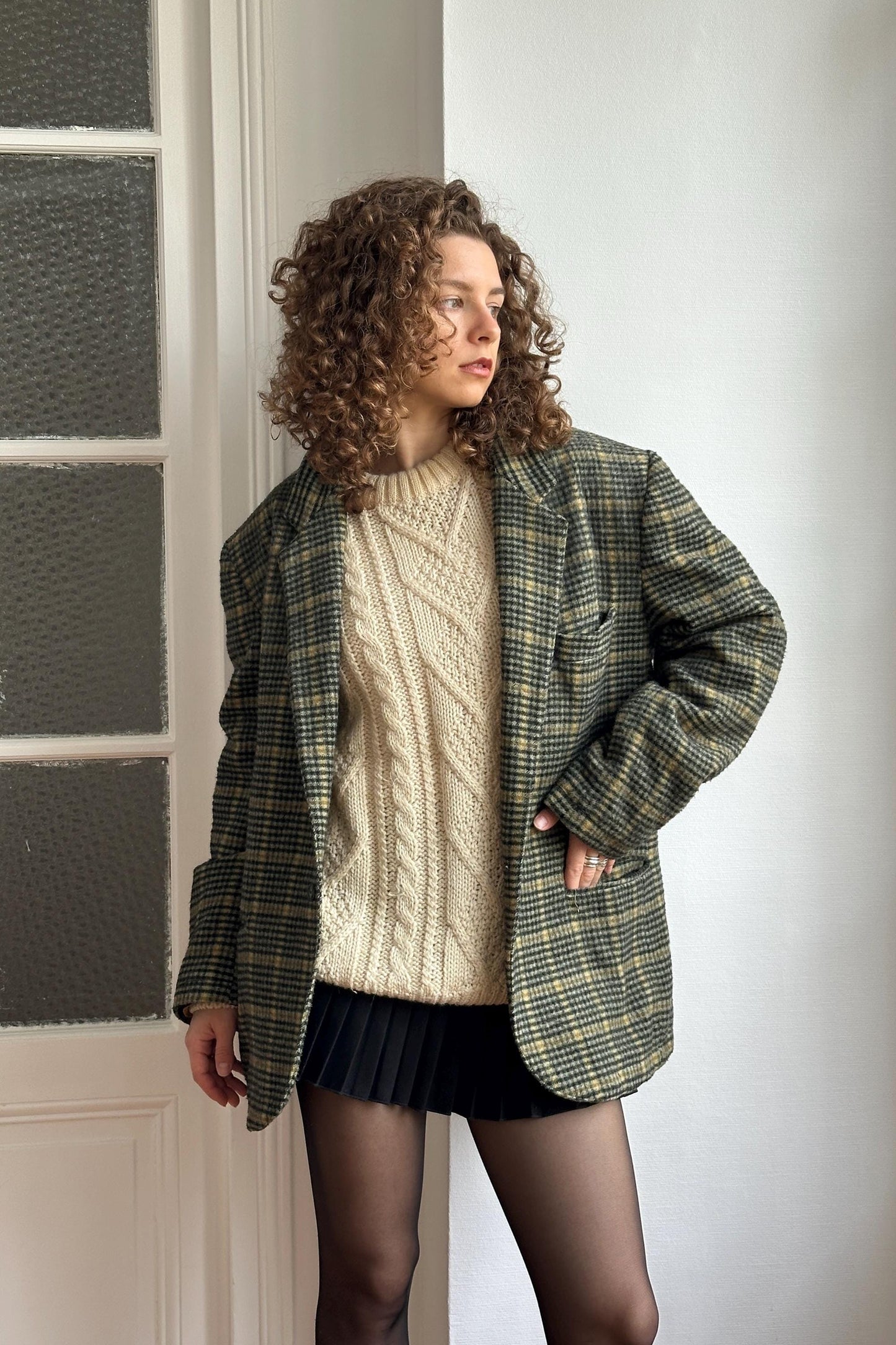 A woman with curly hair wearing a plaid jacket, a white sweater, and a black skirt stands in front of a white door.