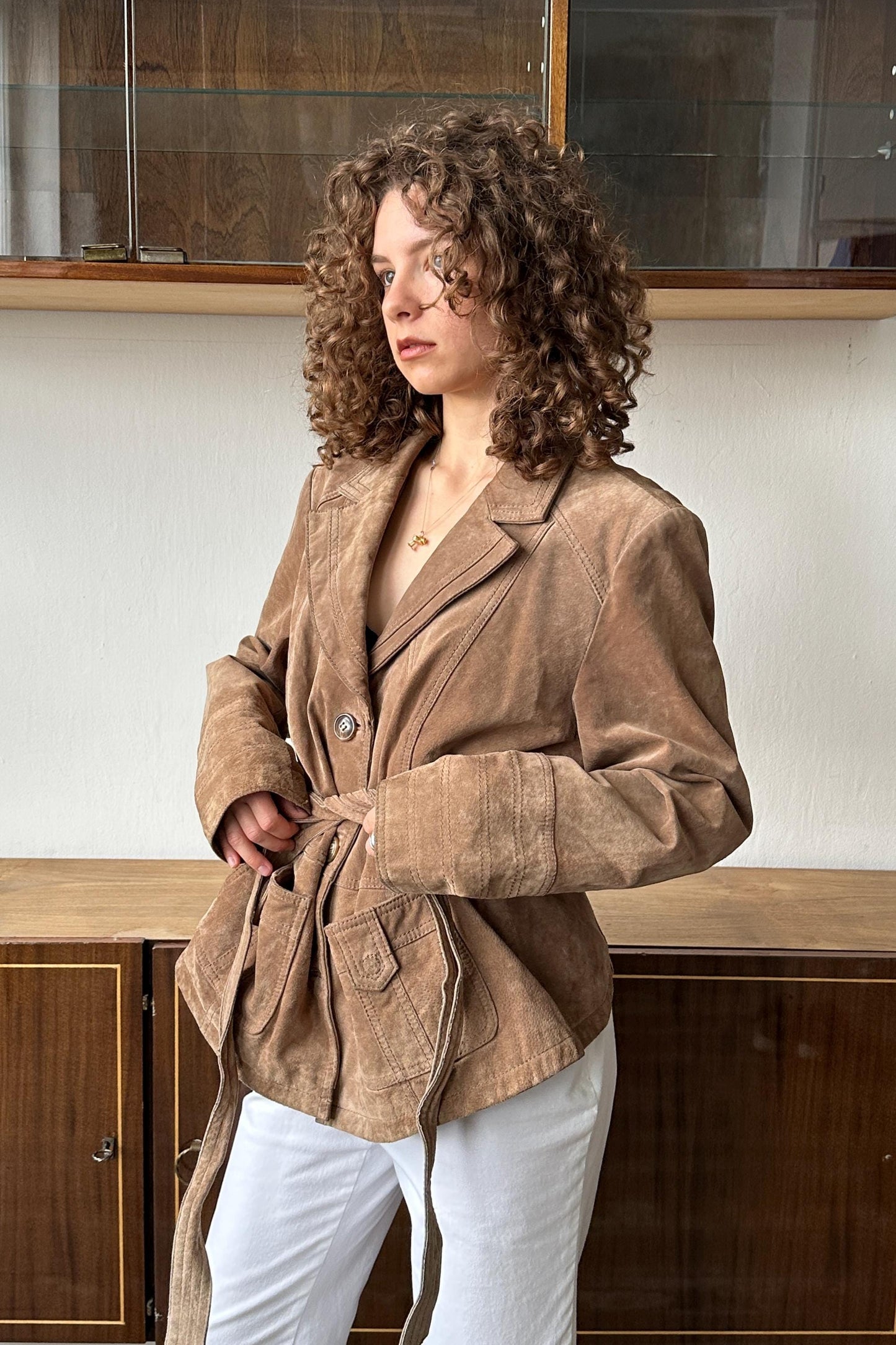 A woman with curly hair wearing a tan jacket stands in front of a wooden cabinet.