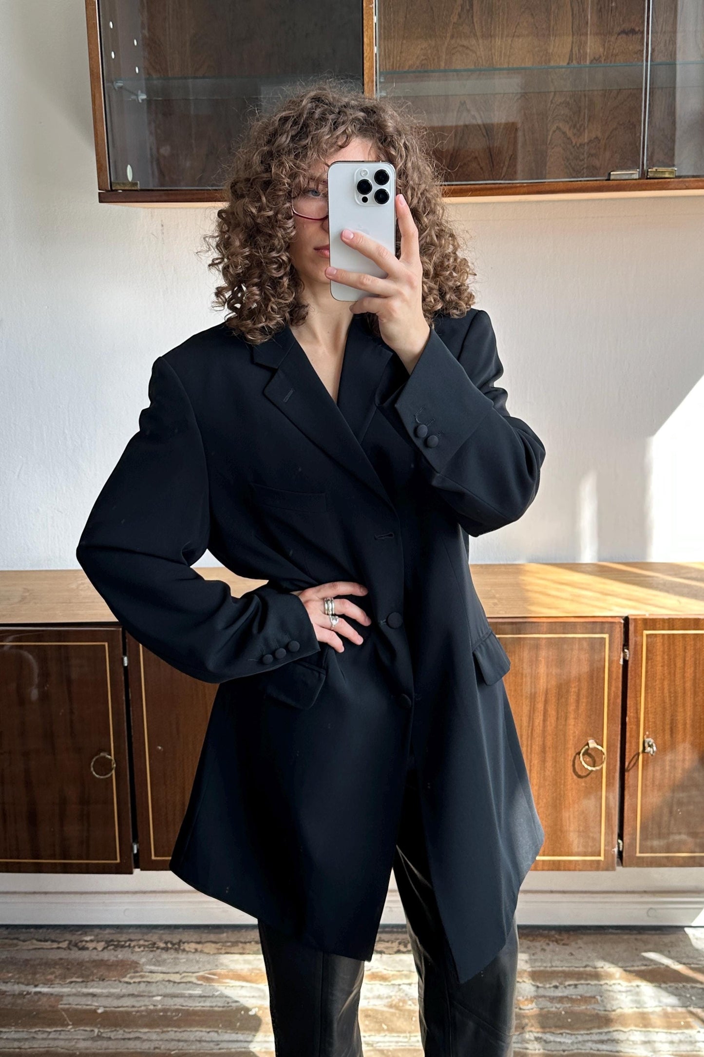 A woman with curly hair is taking a selfie with a white iPhone, wearing a black blazer.