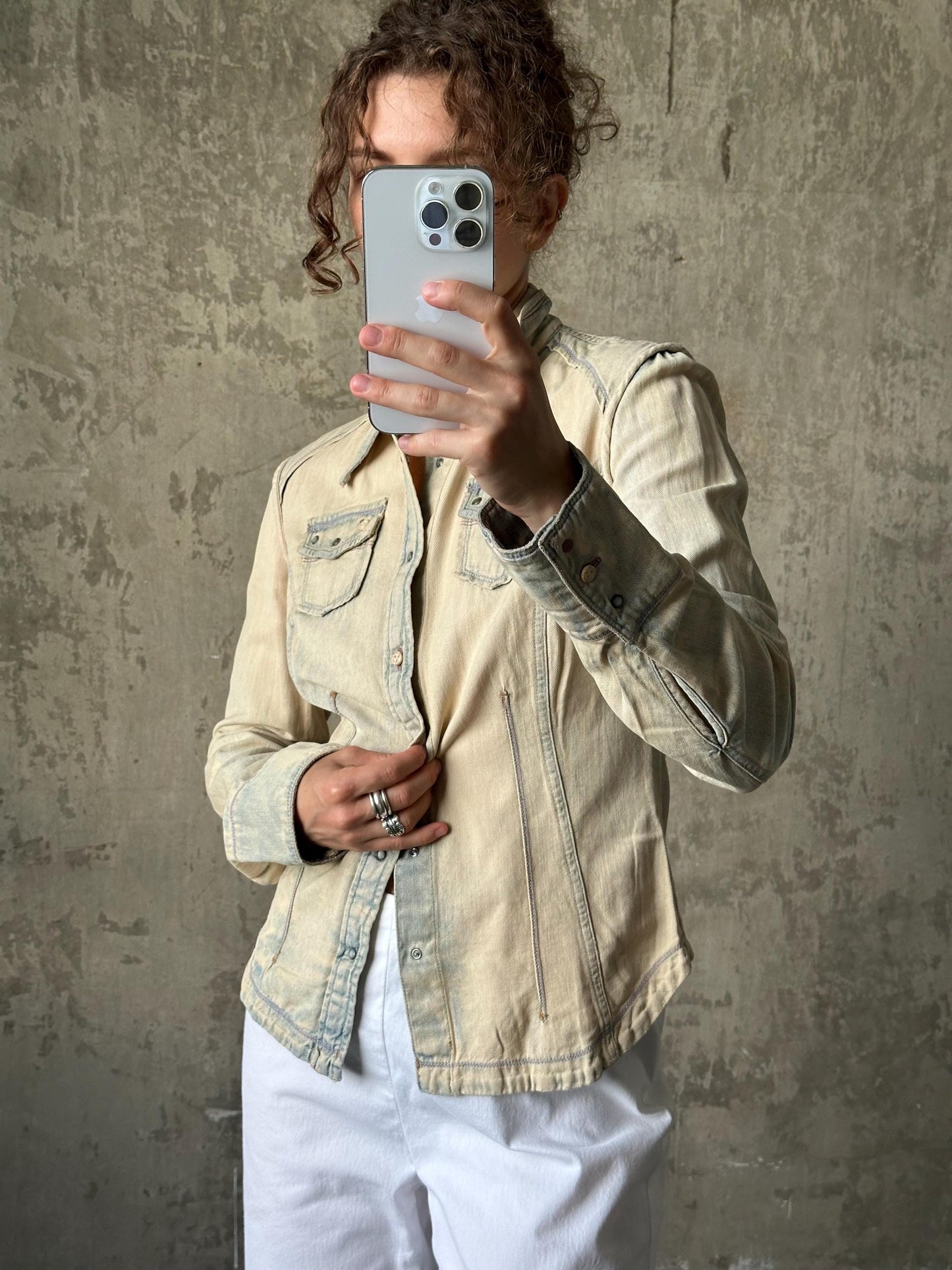 A person is taking a selfie with a smartphone, wearing a beige jacket and white pants against a concrete wall background.