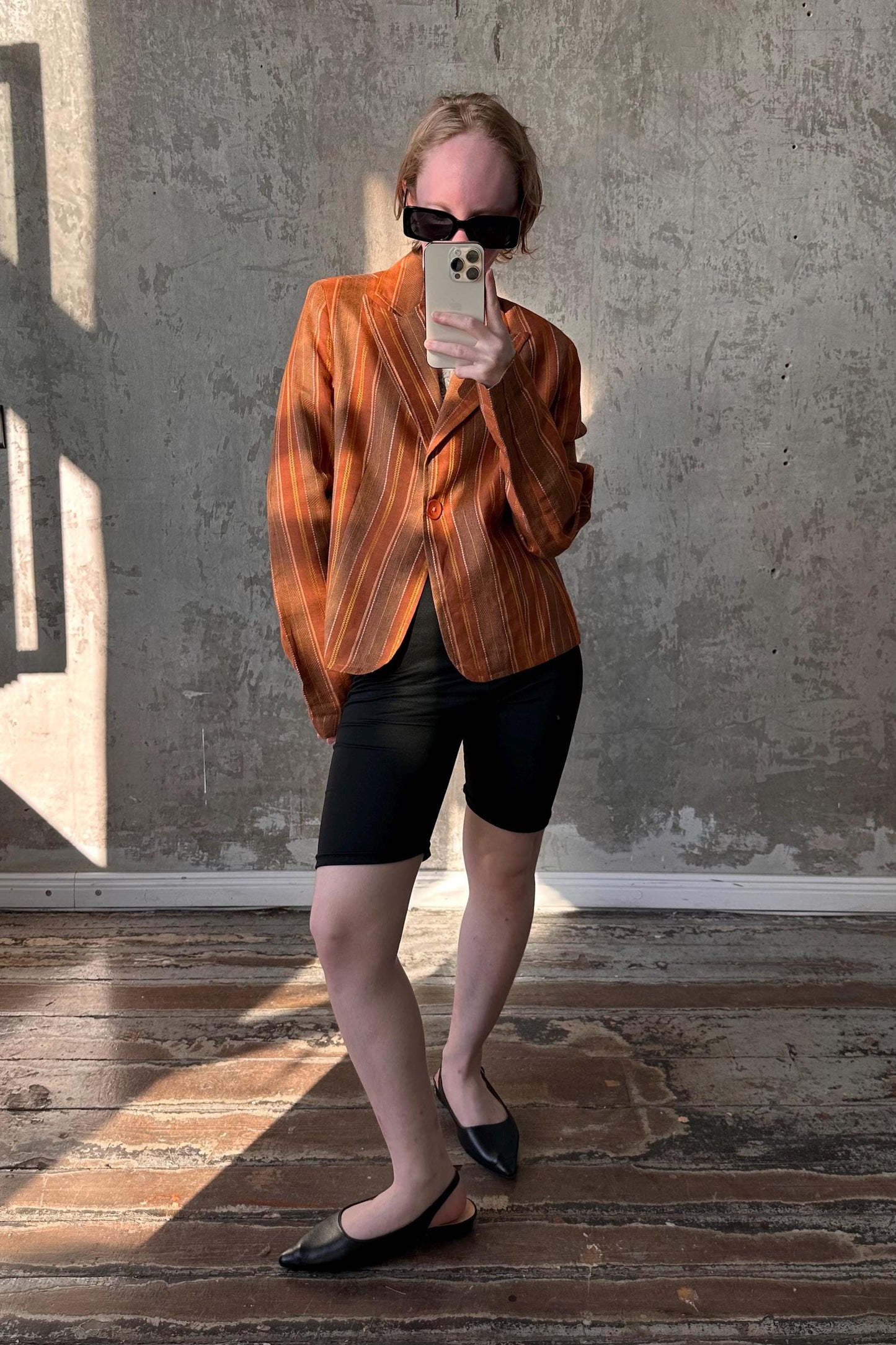 A person is taking a selfie with a smartphone, wearing a striped orange jacket, black shorts, and black shoes, standing in front of a concrete wall.