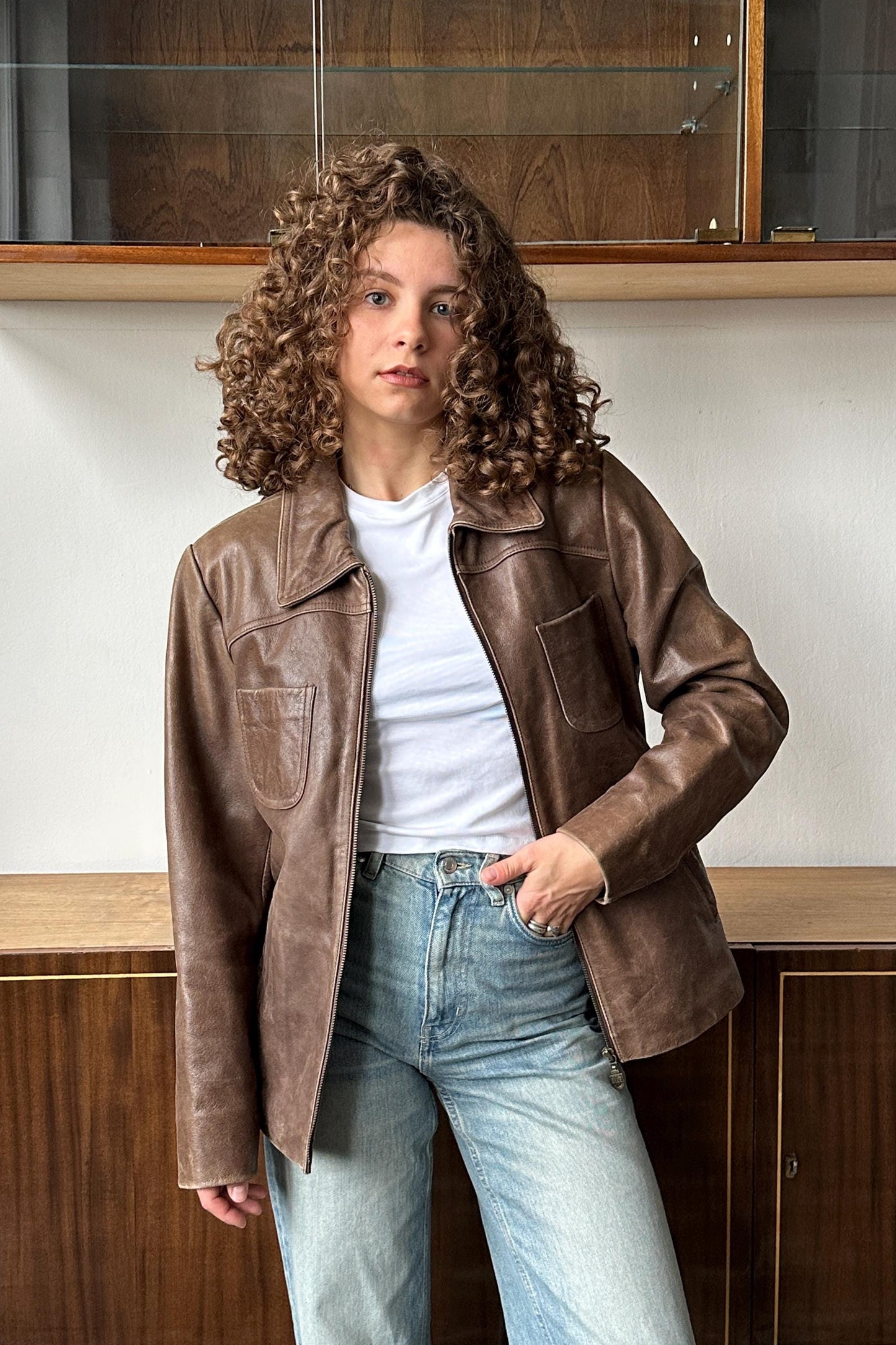 A young woman with curly hair wearing a brown leather jacket and blue jeans stands in front of a wooden cabinet.