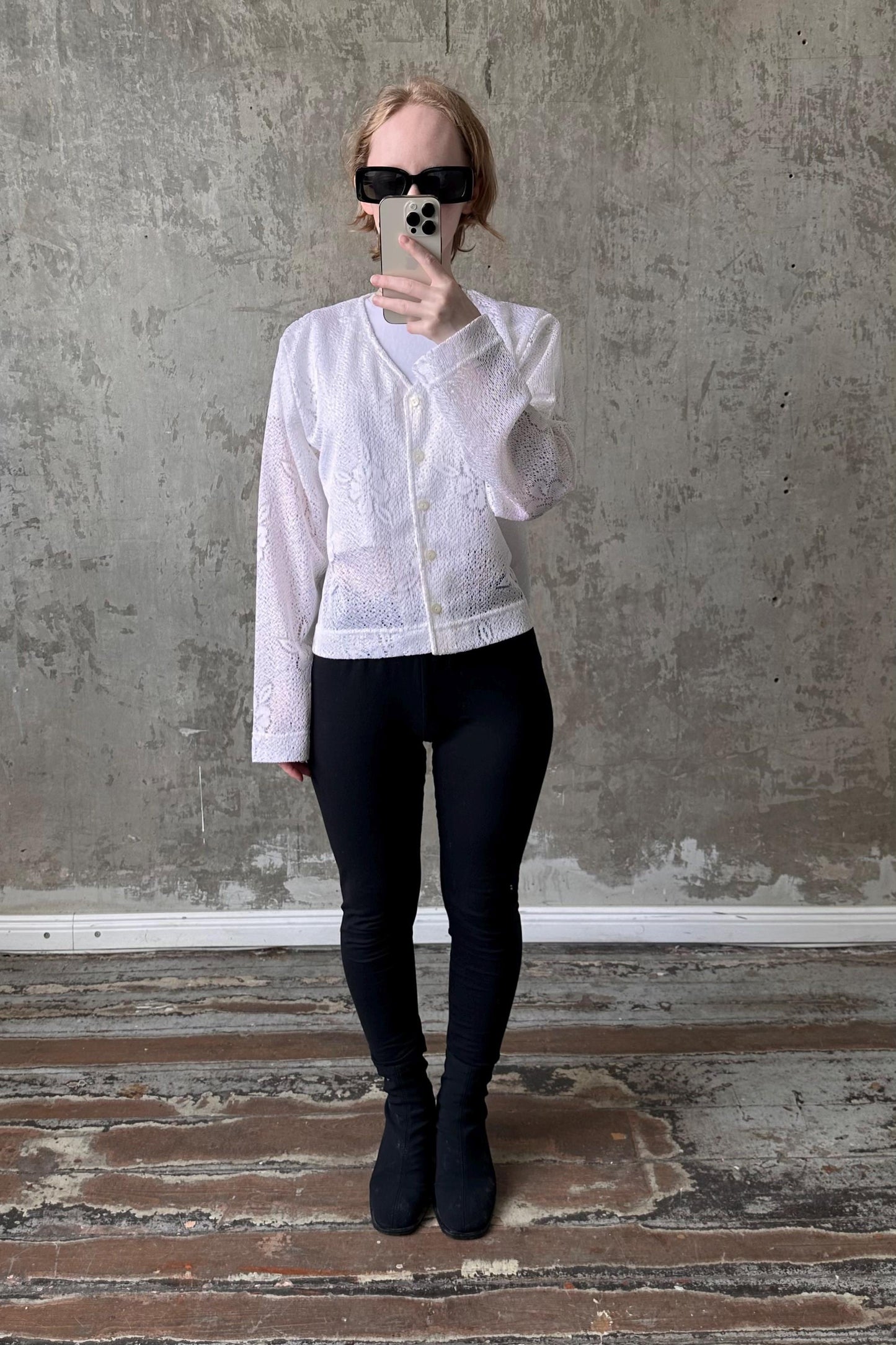 A person is taking a selfie with a white lace top and black leggings, standing in front of a concrete wall.