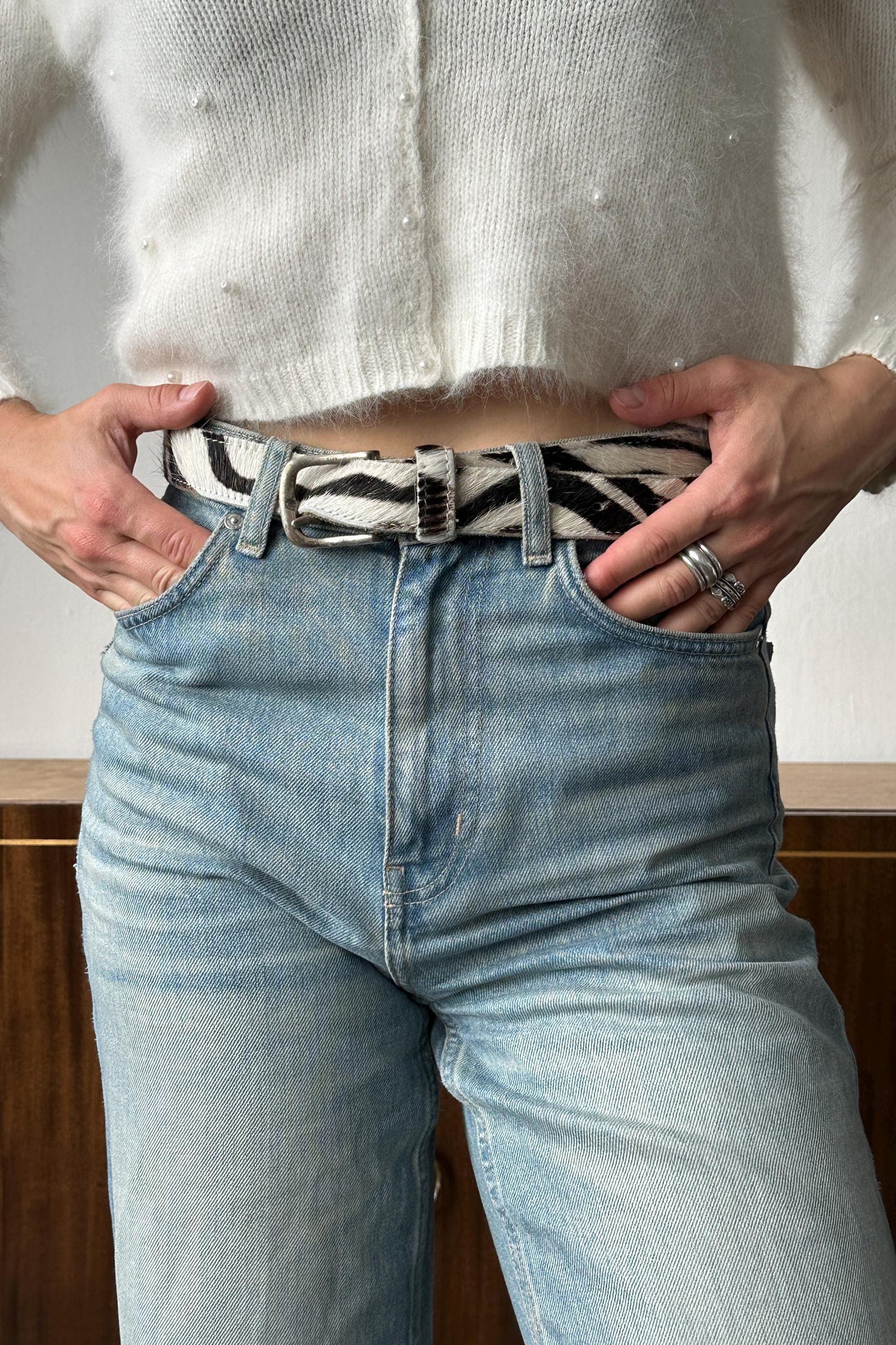 A person wearing a white sweater, blue jeans, and a black and white zebra-striped belt stands with their hands in their pockets.