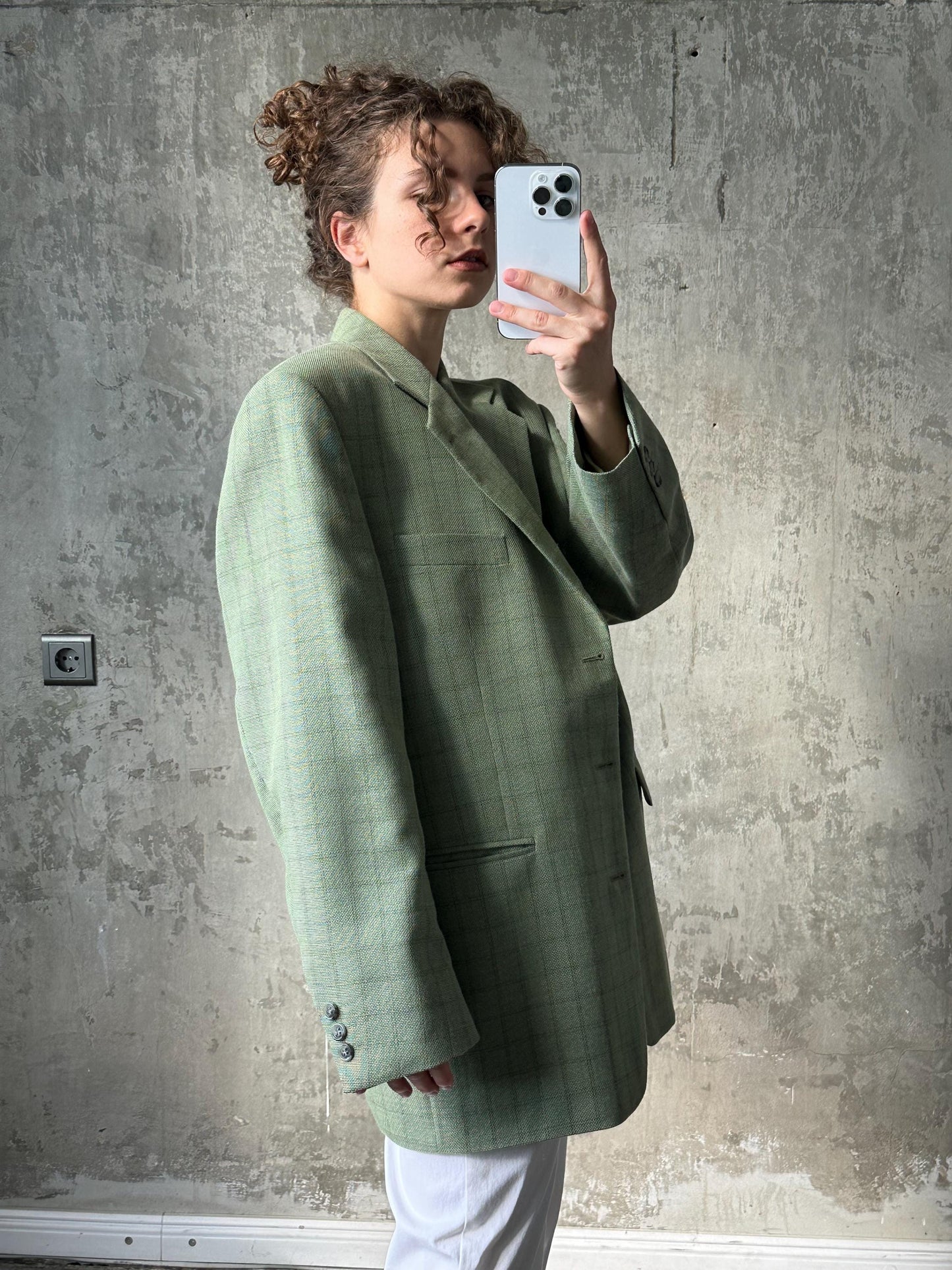 A woman in a green plaid blazer is taking a selfie with a smartphone, capturing her reflection in the mirror.