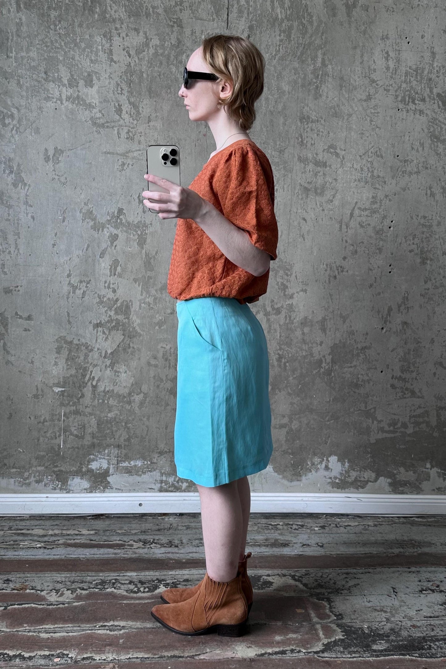 A woman in an orange top and blue skirt stands in front of a concrete wall, holding a phone and wearing sunglasses.
