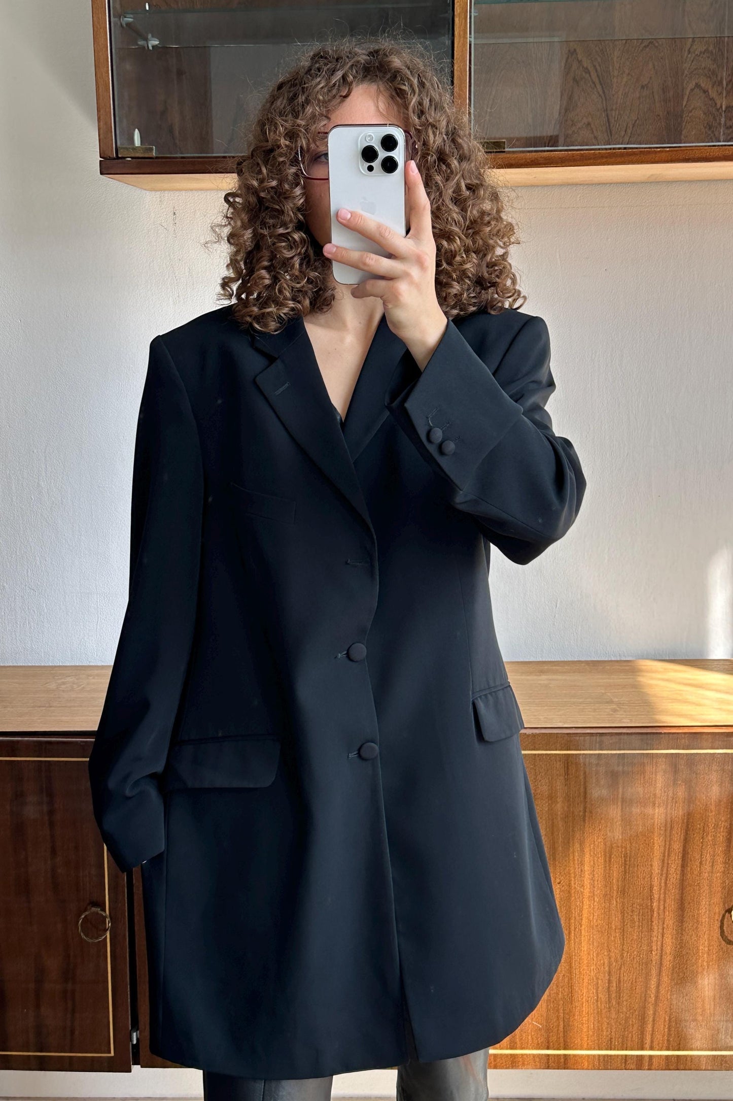 A person with curly hair is taking a selfie with a smartphone, wearing a black blazer.