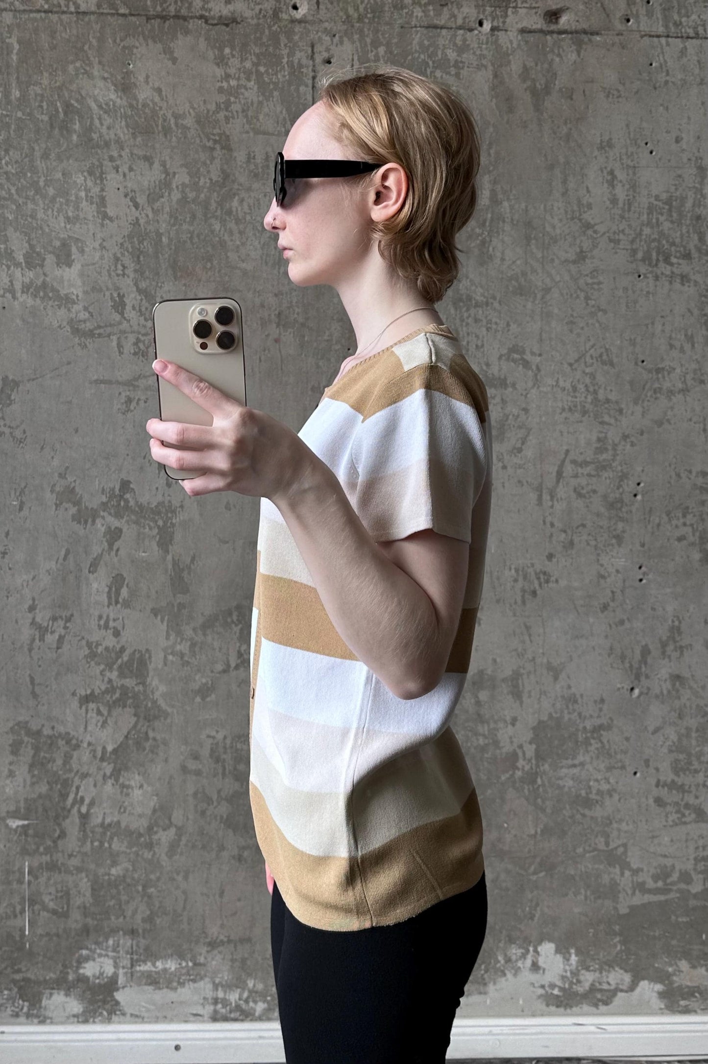 A woman wearing sunglasses is holding a smartphone and taking a selfie in front of a concrete wall.