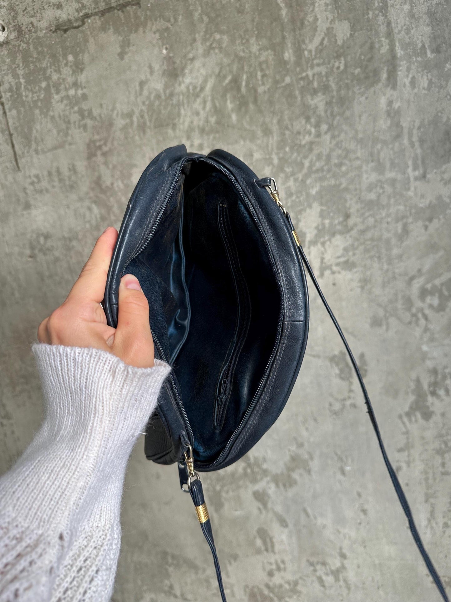 A hand holding a black leather crossbody bag against a concrete wall background.