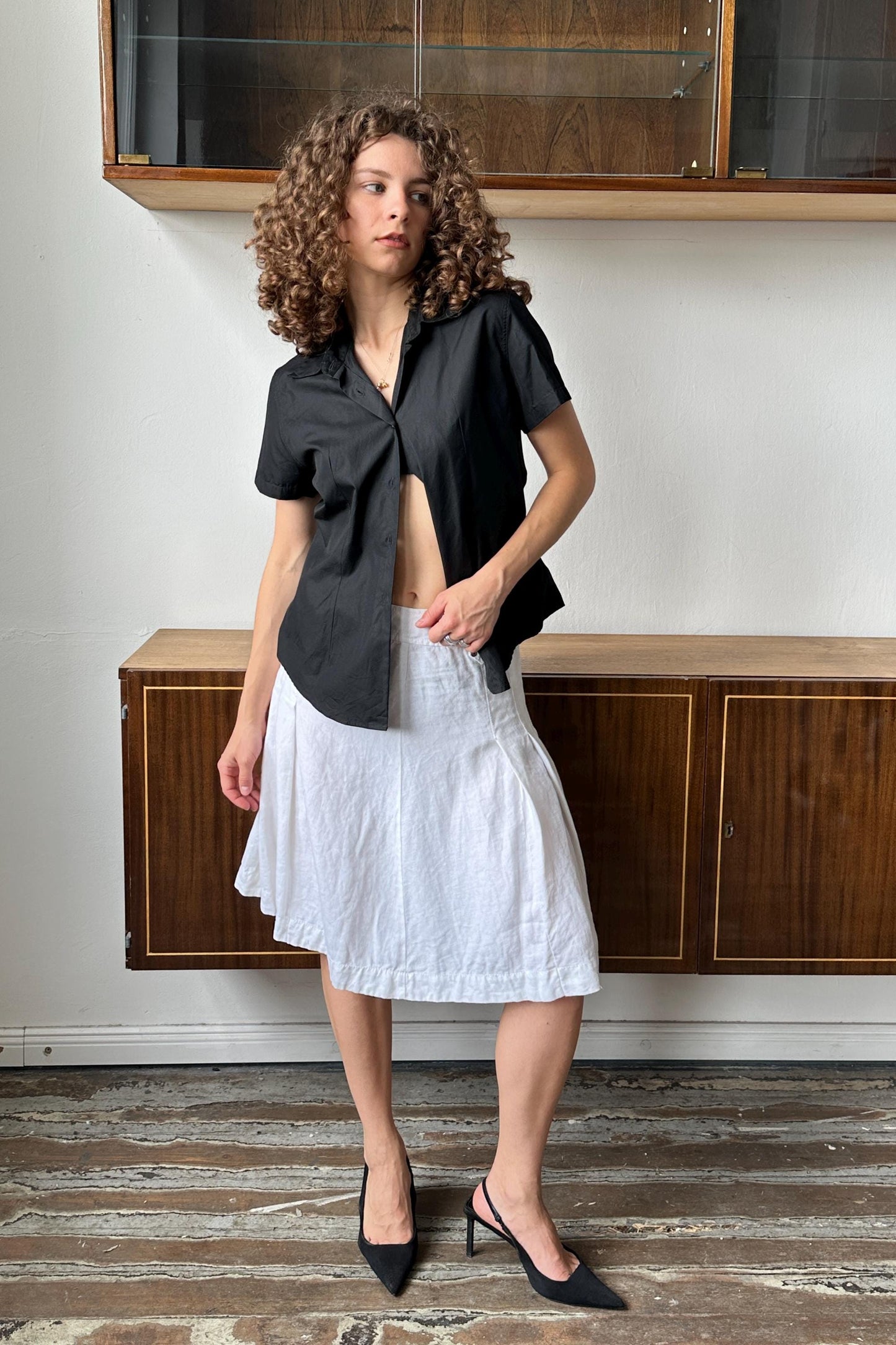 A woman with curly hair wearing a black shirt and white skirt stands in a room with wooden cabinets in the background.