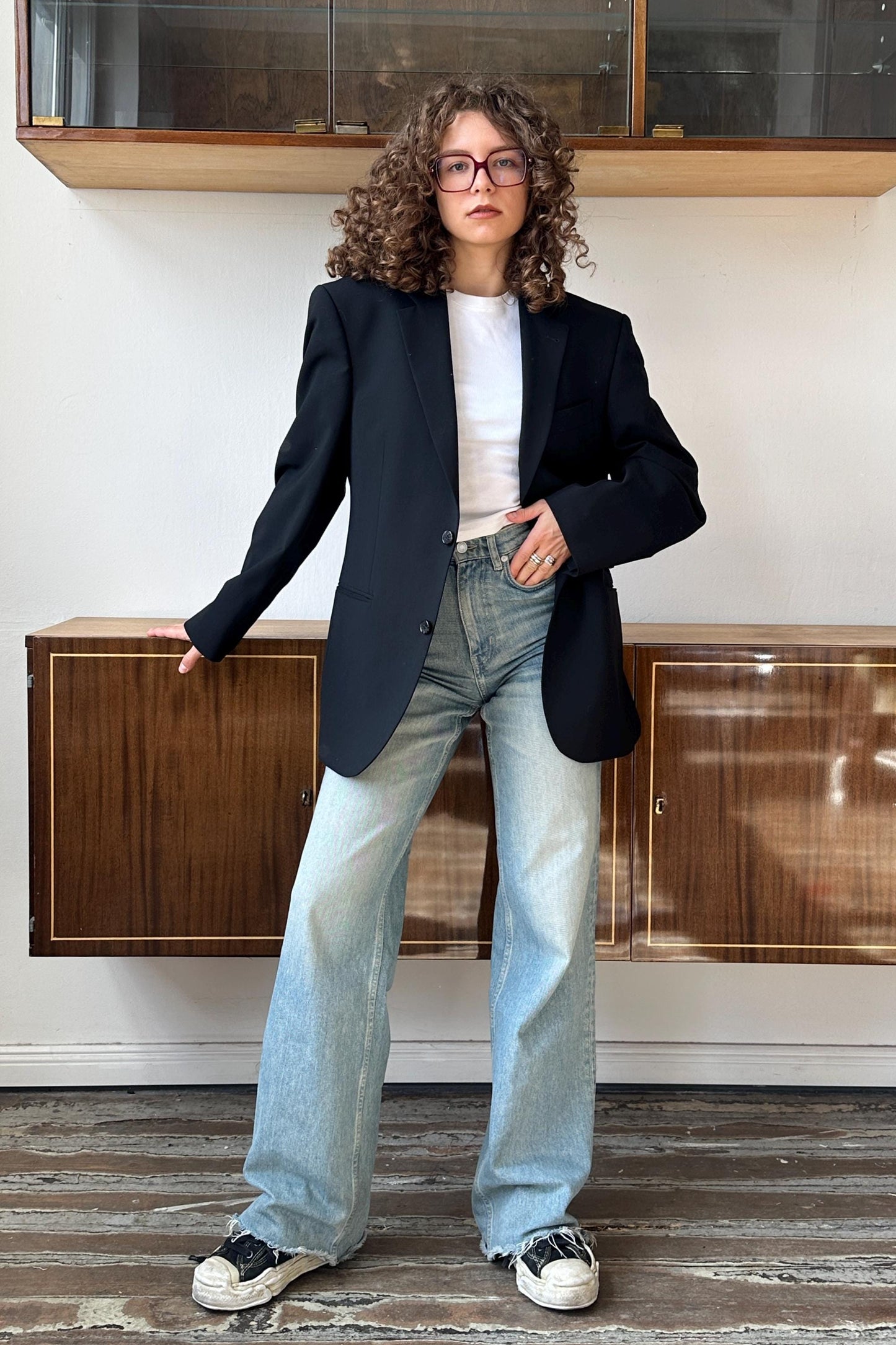 A woman with curly hair wearing a black blazer, white shirt, and blue jeans stands in front of a wooden cabinet.