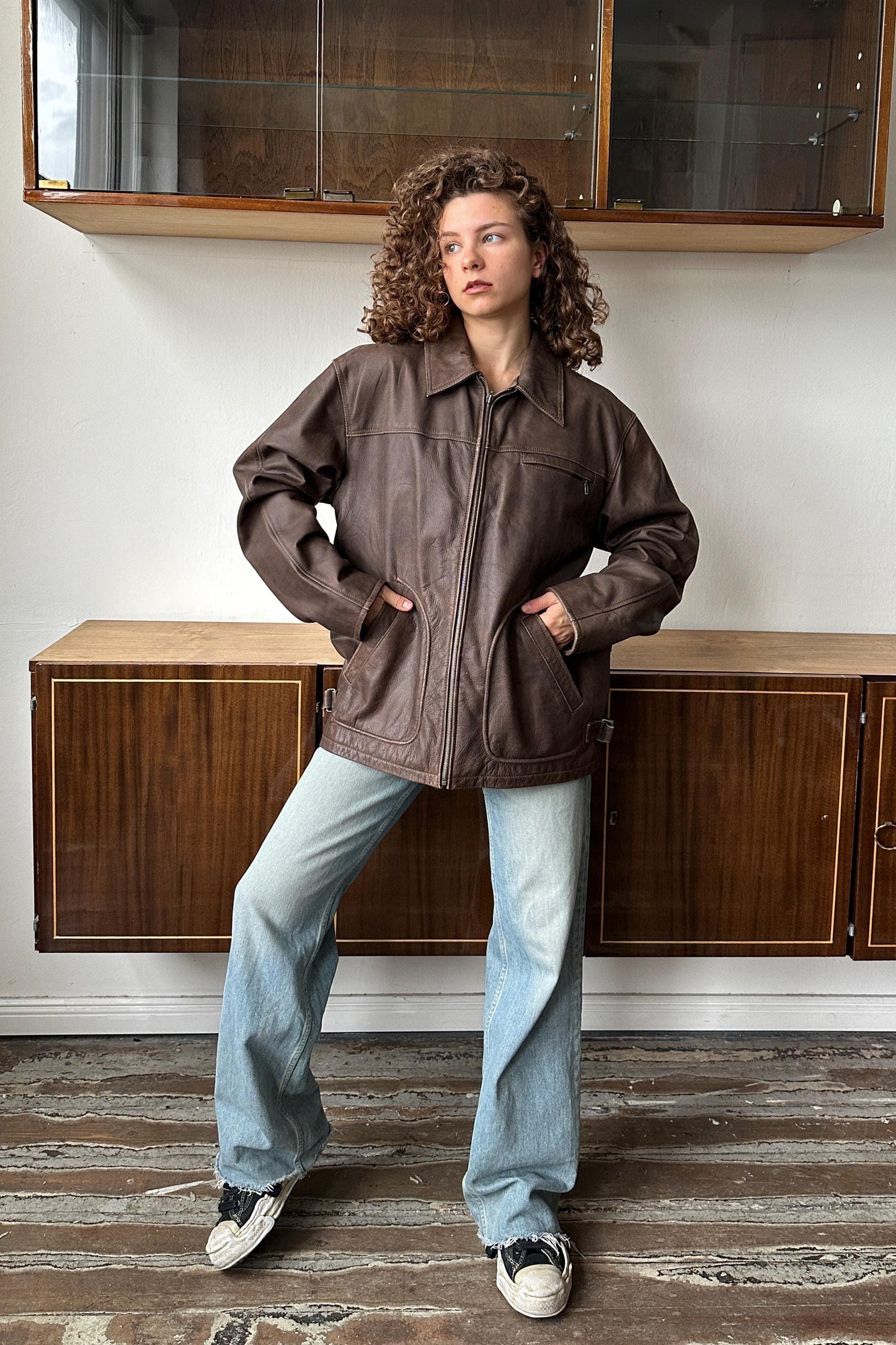 A young woman with curly hair stands in a room, wearing a brown leather jacket and blue jeans.