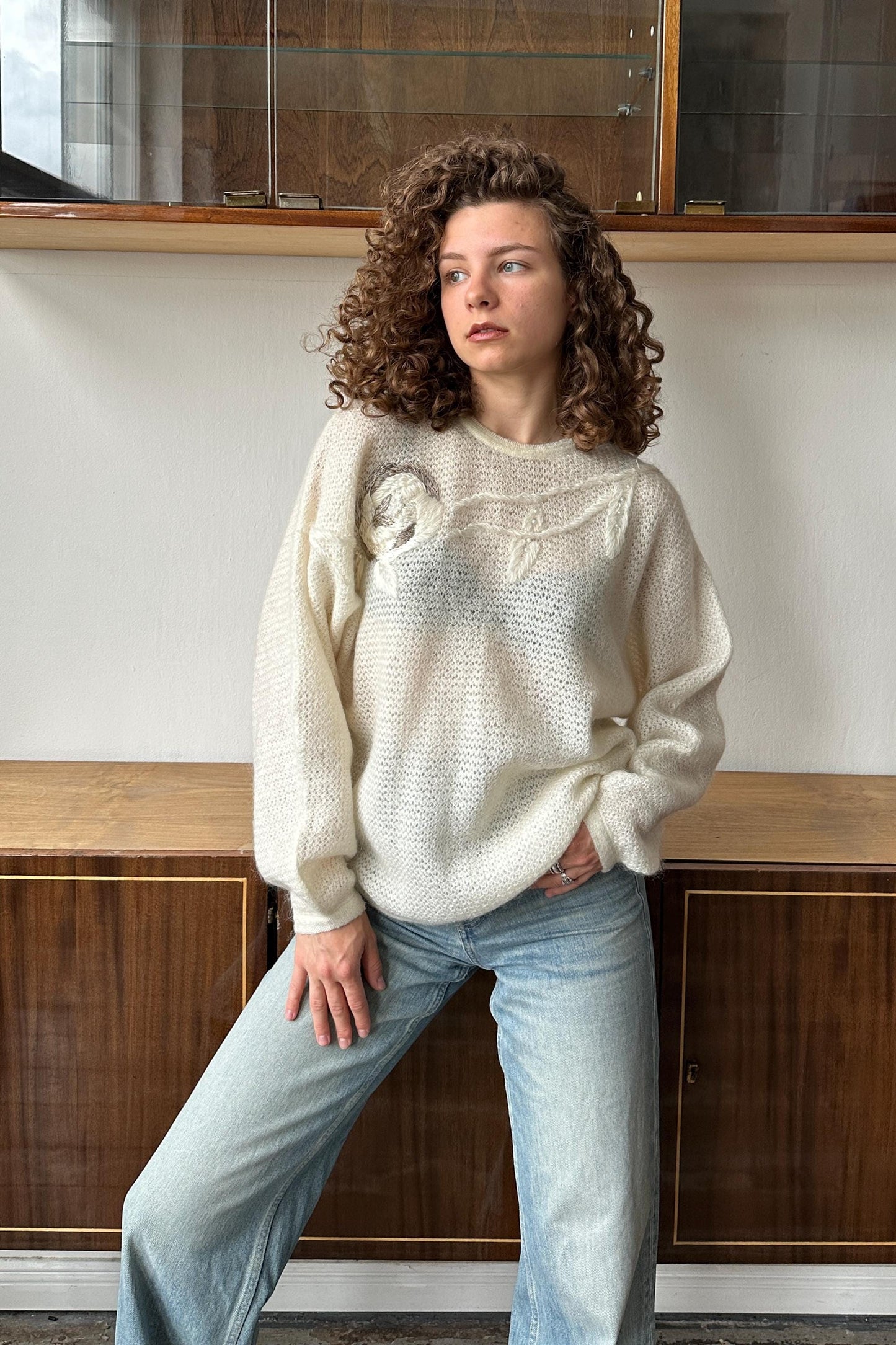 A woman with curly hair wearing a white sweater and blue jeans stands in front of a wooden cabinet.