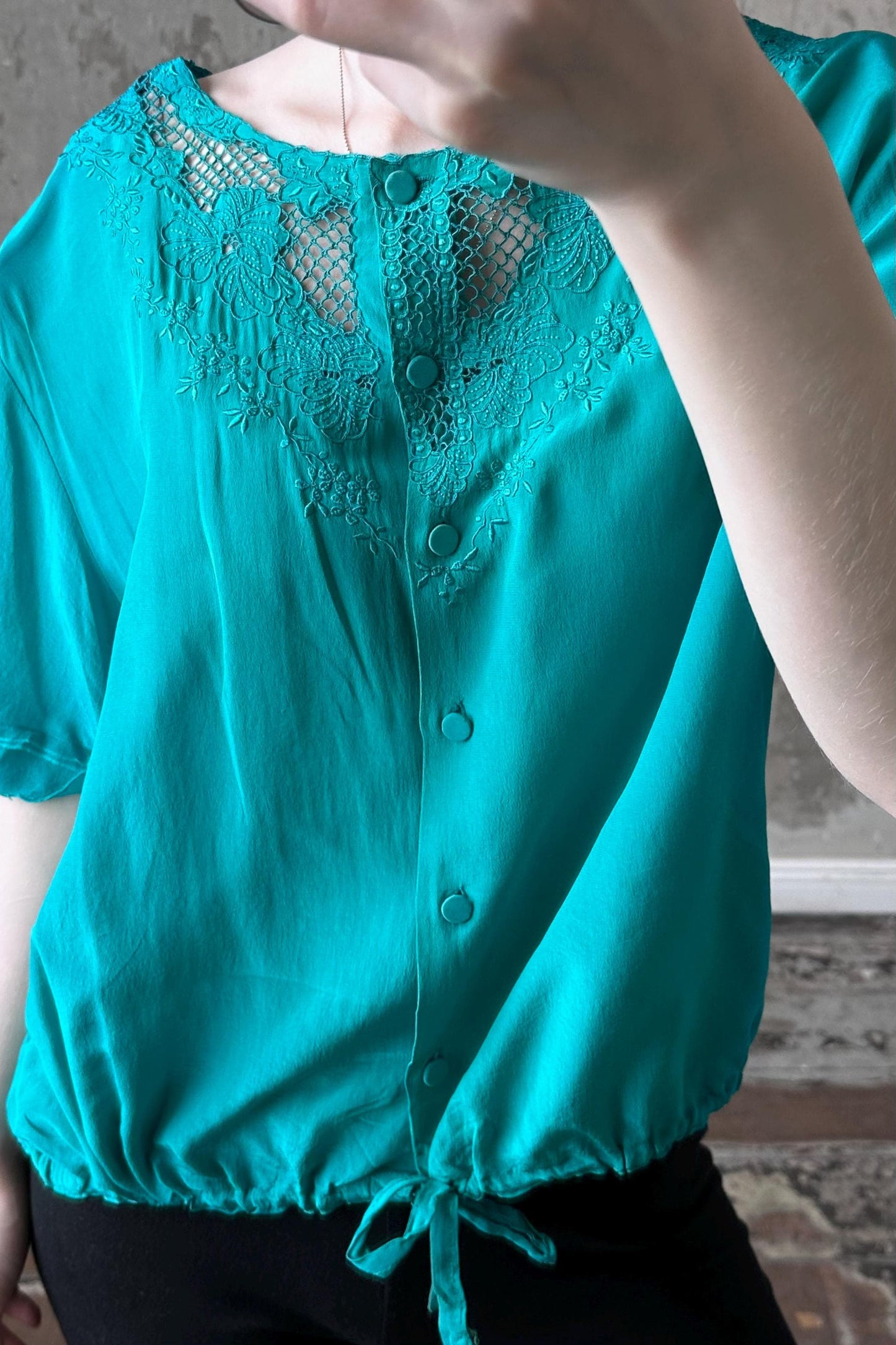A person wearing a turquoise blouse with lace detailing and buttons, standing in front of a concrete wall.