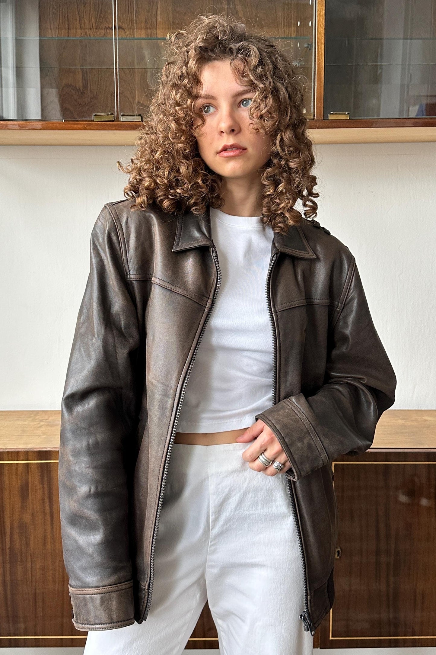 A woman with curly hair wearing a brown leather jacket and white pants stands in front of a wooden cabinet.