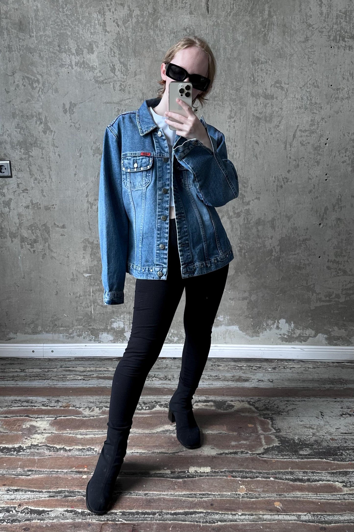 A person wearing a denim jacket, black pants, and sunglasses is taking a selfie with a smartphone in front of a concrete wall.