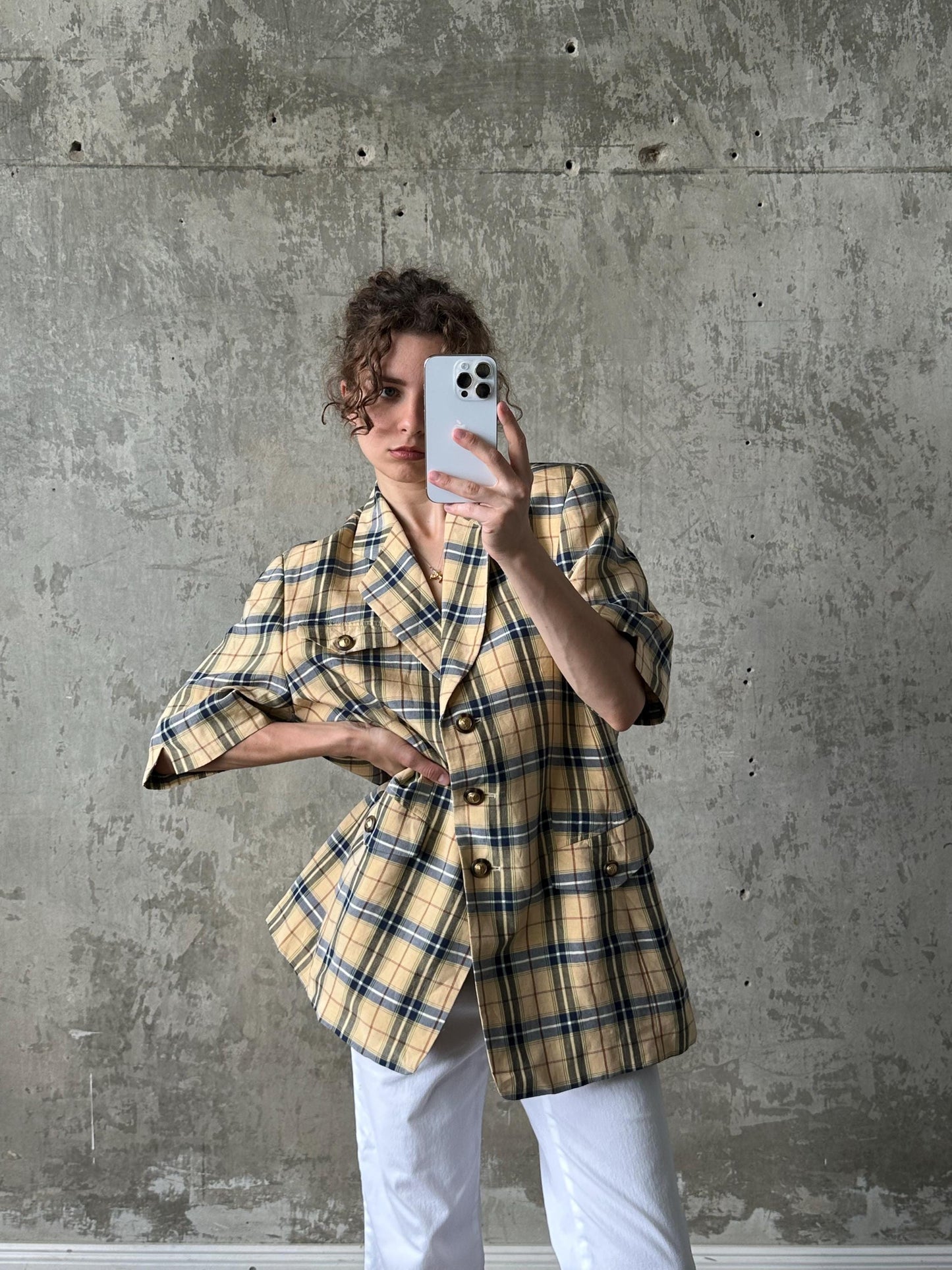 A woman wearing a yellow and black plaid blazer is taking a selfie with a white phone against a concrete wall.