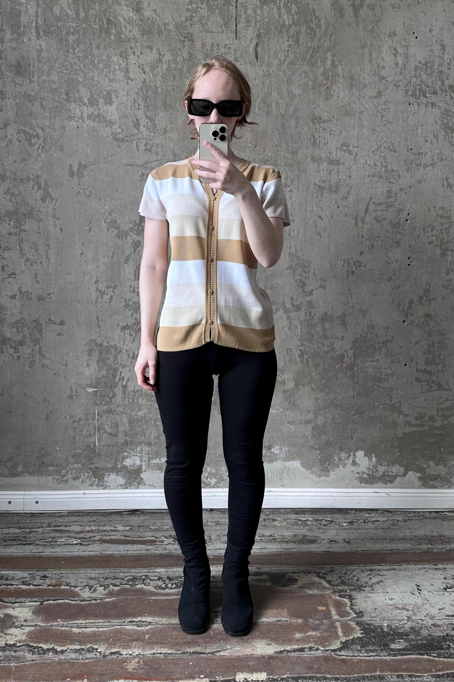 A woman wearing sunglasses and a striped sweater is taking a selfie with her phone against a concrete wall.