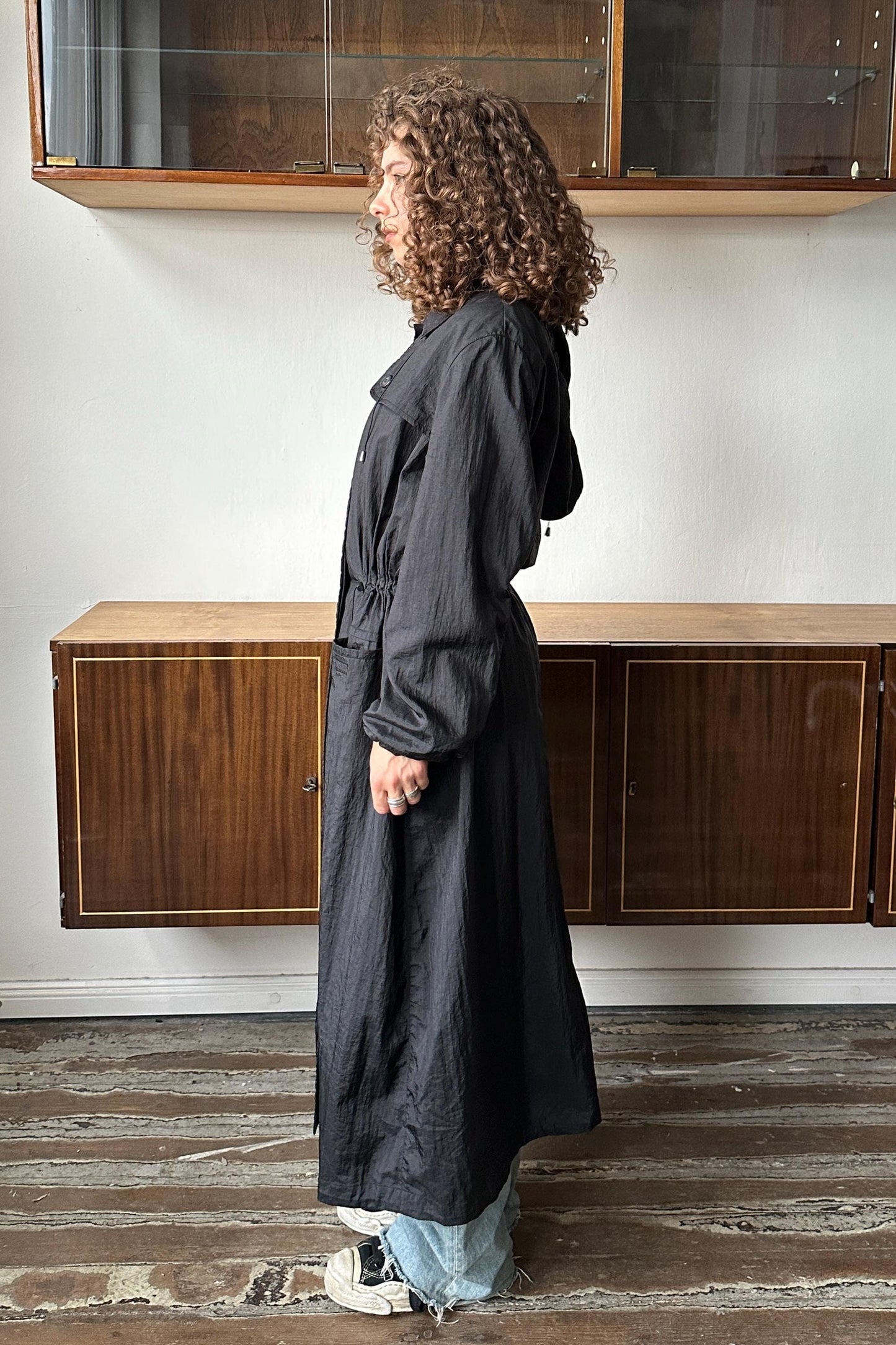 A person with curly hair wearing a long black coat stands in a room with wooden cabinets and a glass door.