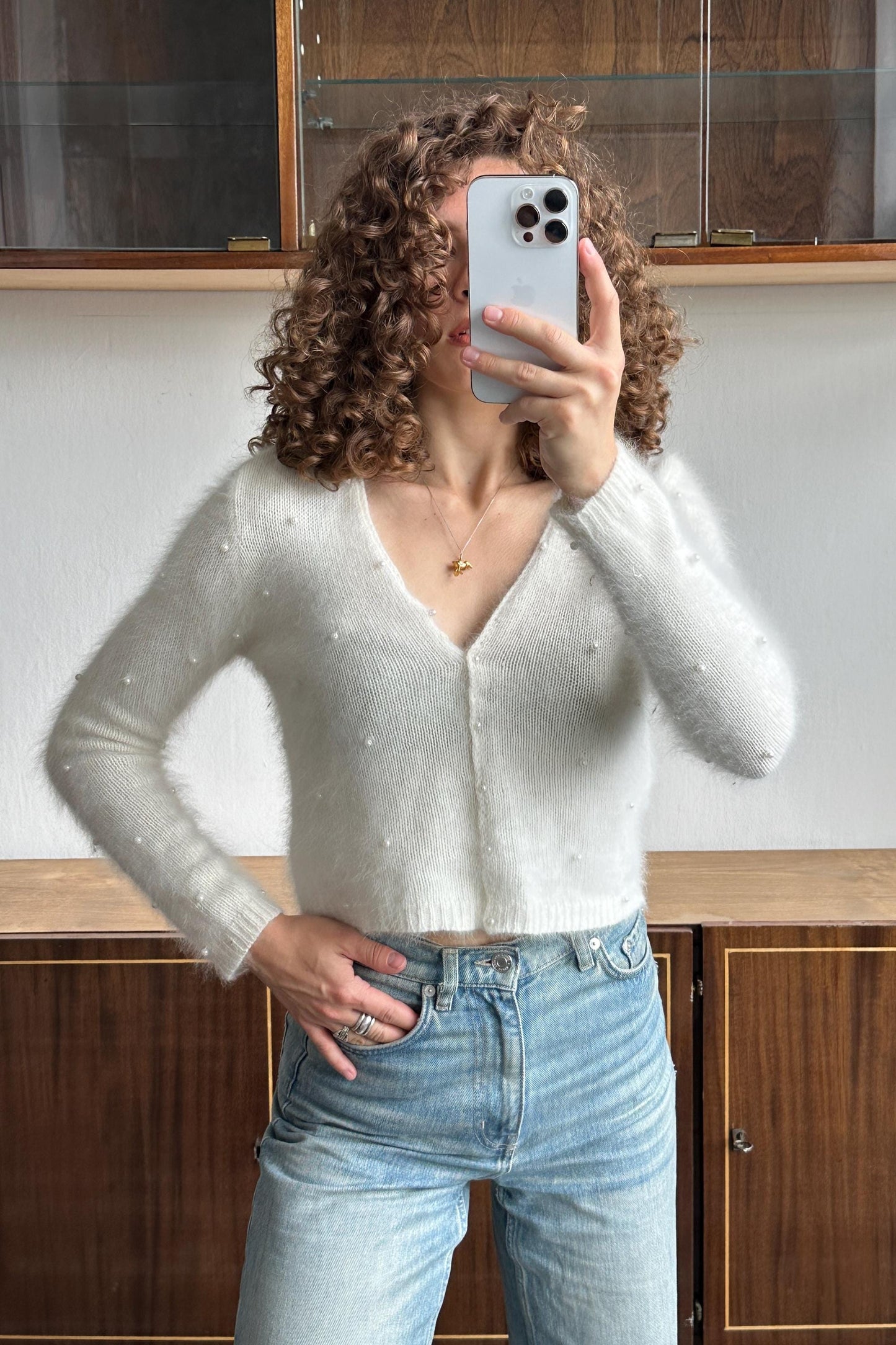 A woman with curly hair is taking a selfie with her iPhone, wearing a white sweater and blue jeans.