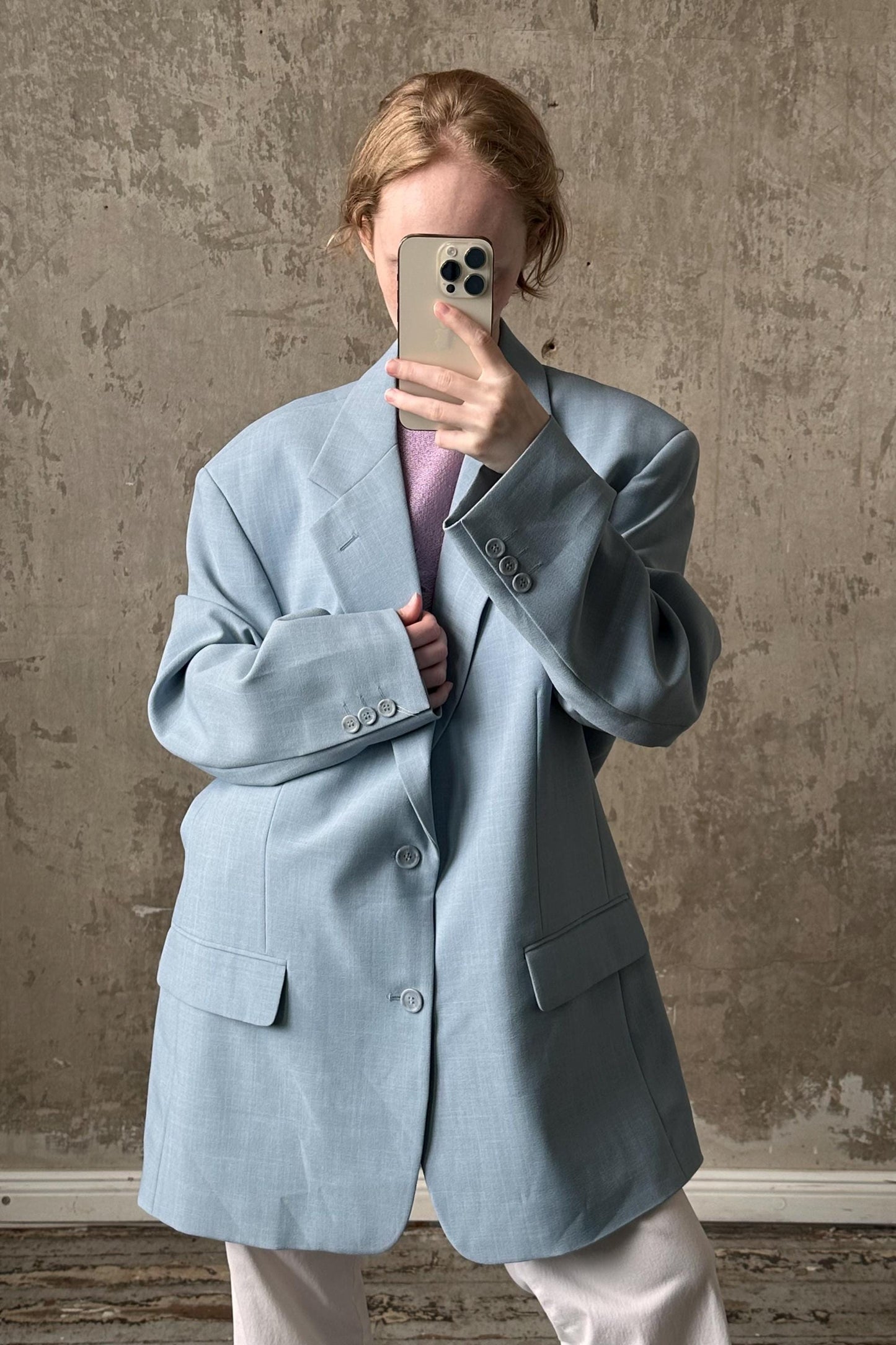 A person is taking a selfie with a smartphone, wearing a light blue blazer and standing in front of a concrete wall.