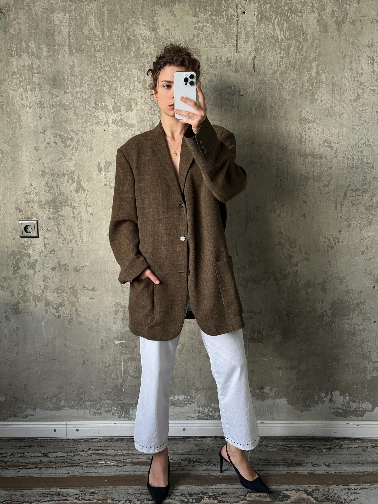 A woman in a brown blazer and white pants is taking a selfie with a white phone against a concrete wall.