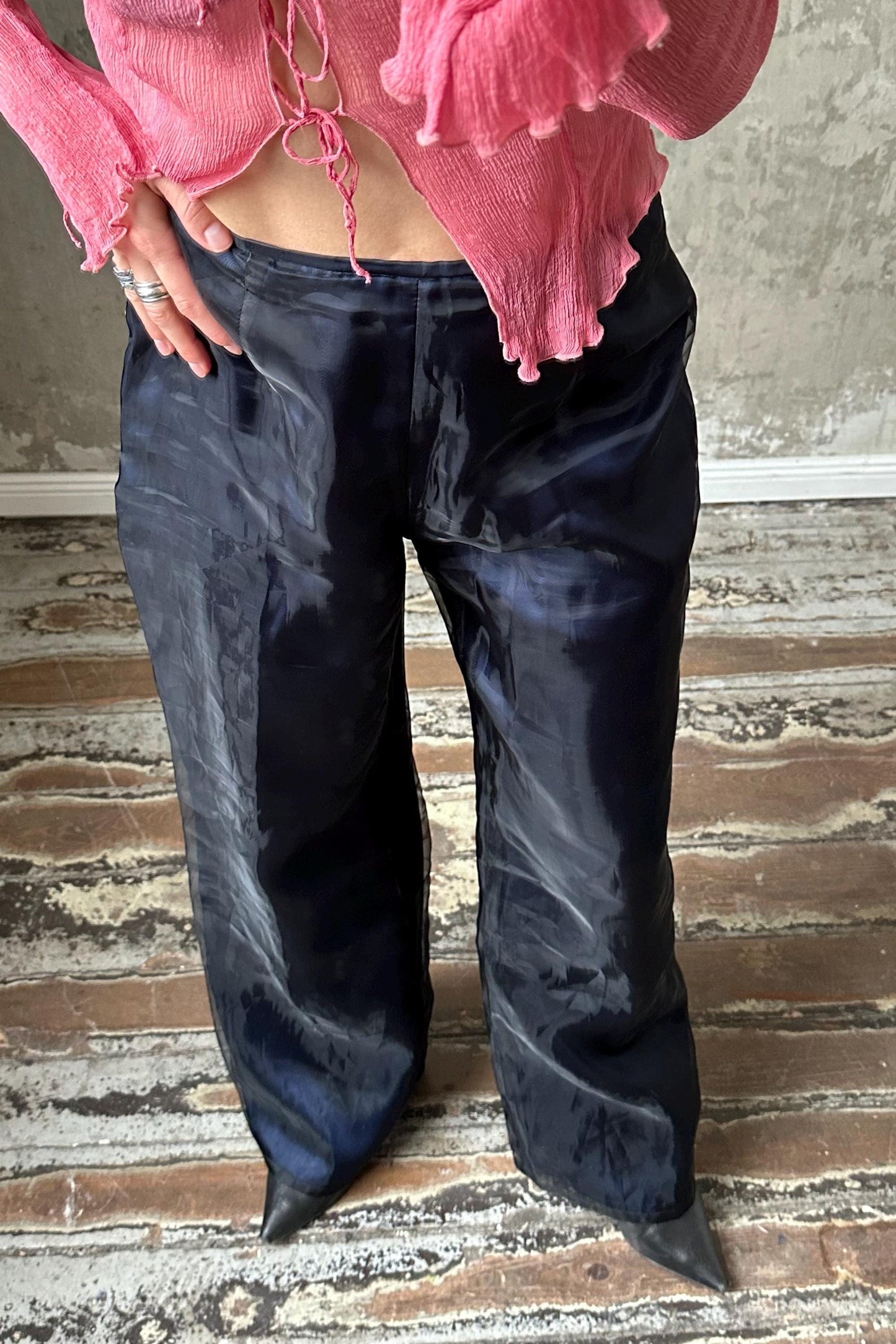 A person wearing a pink blouse and black satin pants stands on a wooden floor.