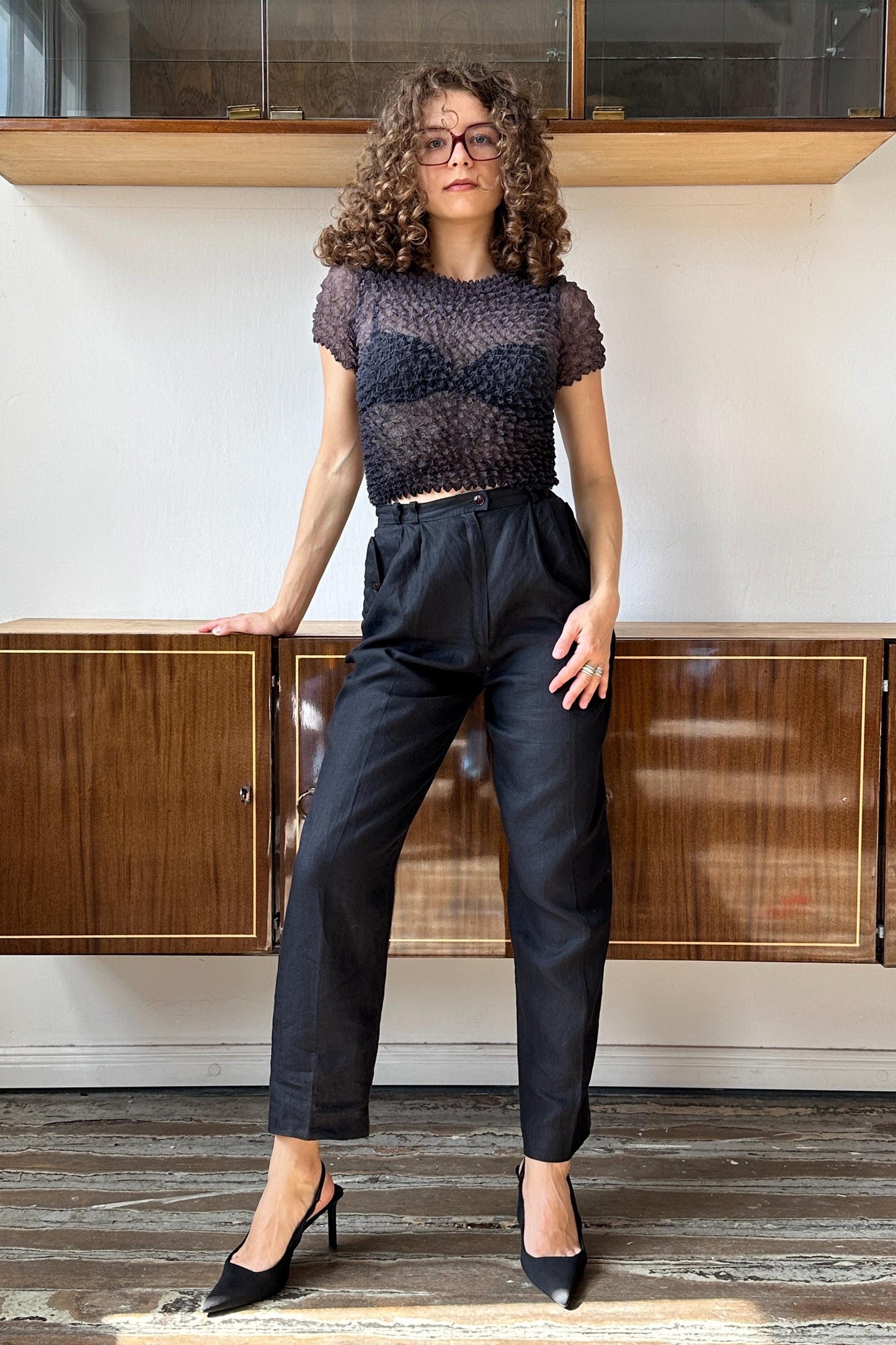 A woman with curly hair and glasses is standing in front of a wooden cabinet, wearing a black top and black pants.