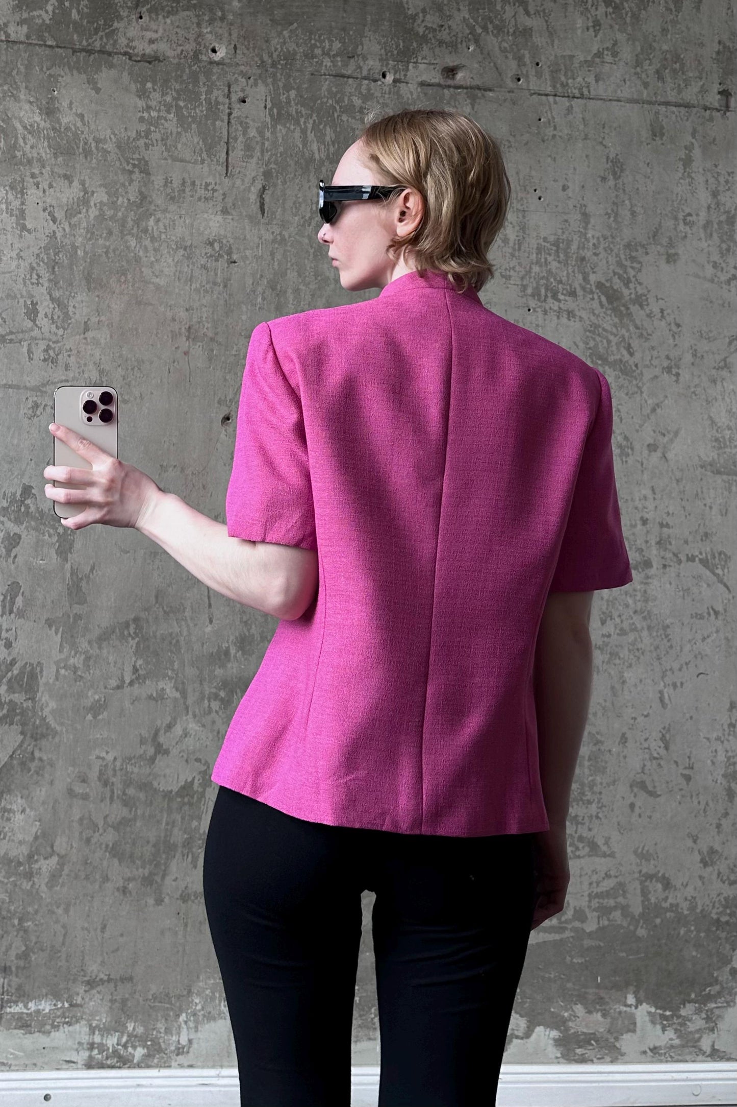 A woman wearing a pink blazer and sunglasses is holding a smartphone in front of a concrete wall.