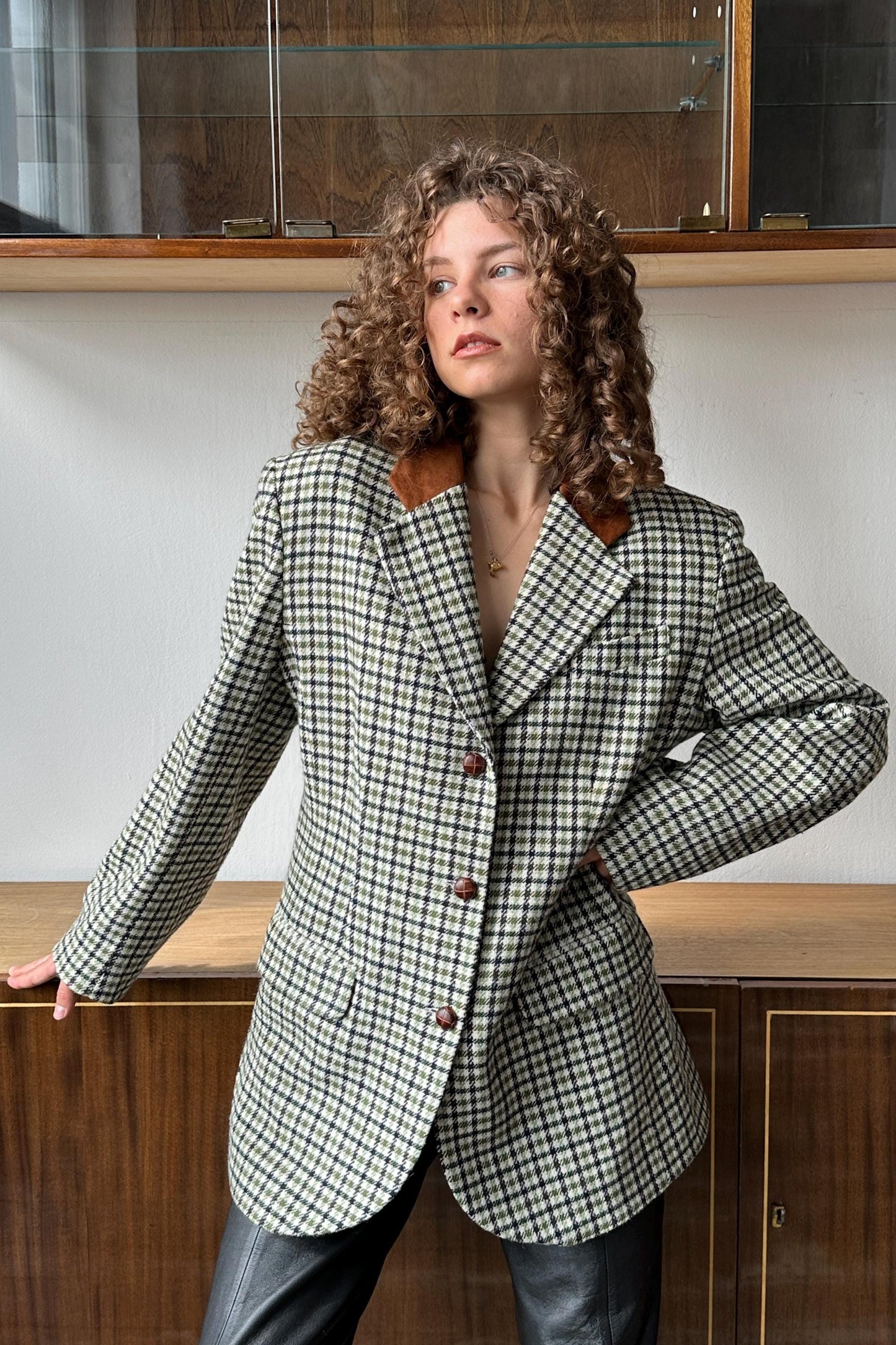 A woman with curly hair wearing a checkered jacket stands in front of a wooden cabinet.