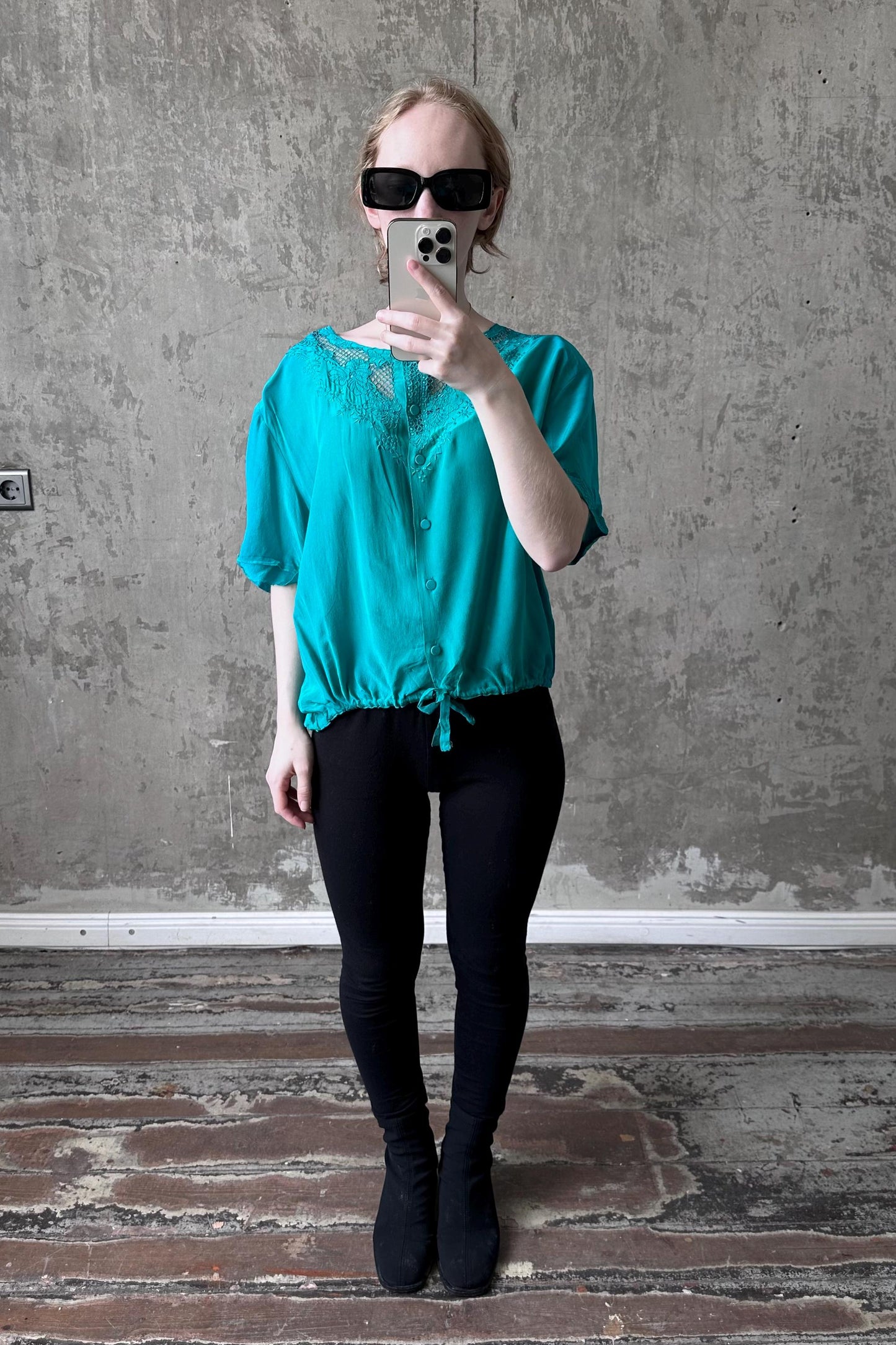 A woman wearing a turquoise blouse and black leggings is taking a selfie with a smartphone, standing in front of a concrete wall.
