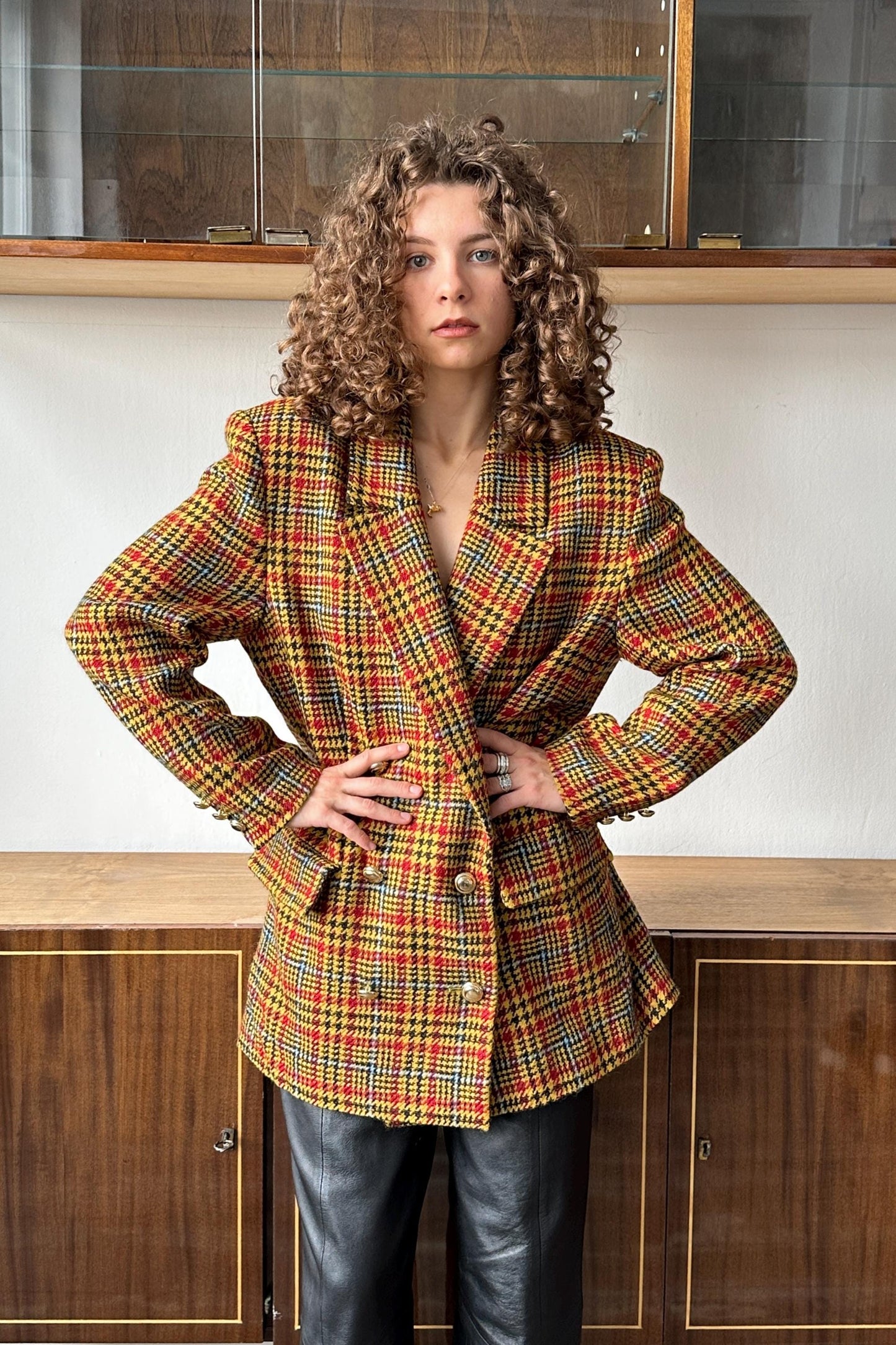 A woman with curly hair wearing a colorful, patterned jacket stands confidently with her hands on her hips.