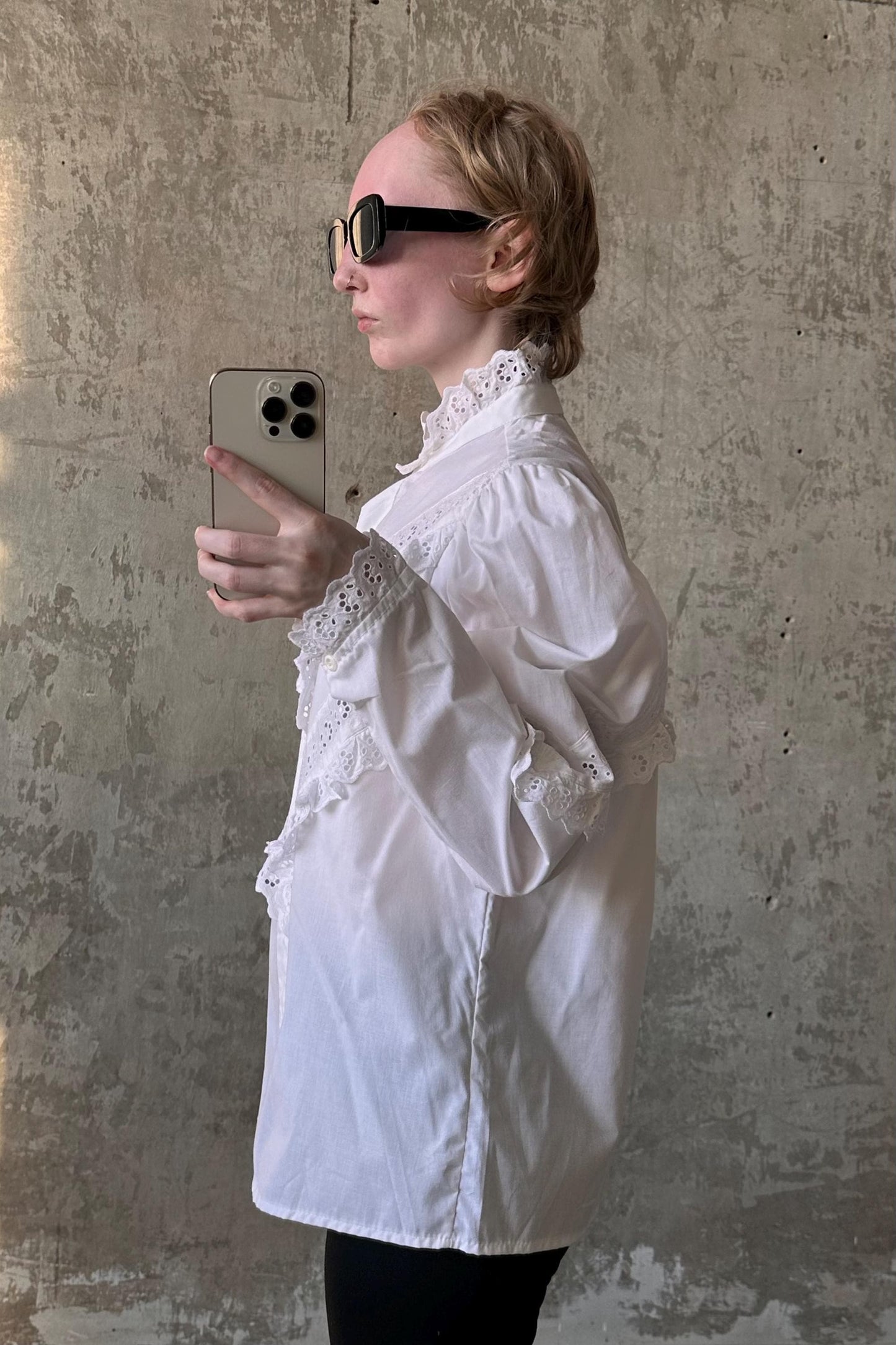 A woman wearing a white blouse and sunglasses is taking a selfie with a smartphone.