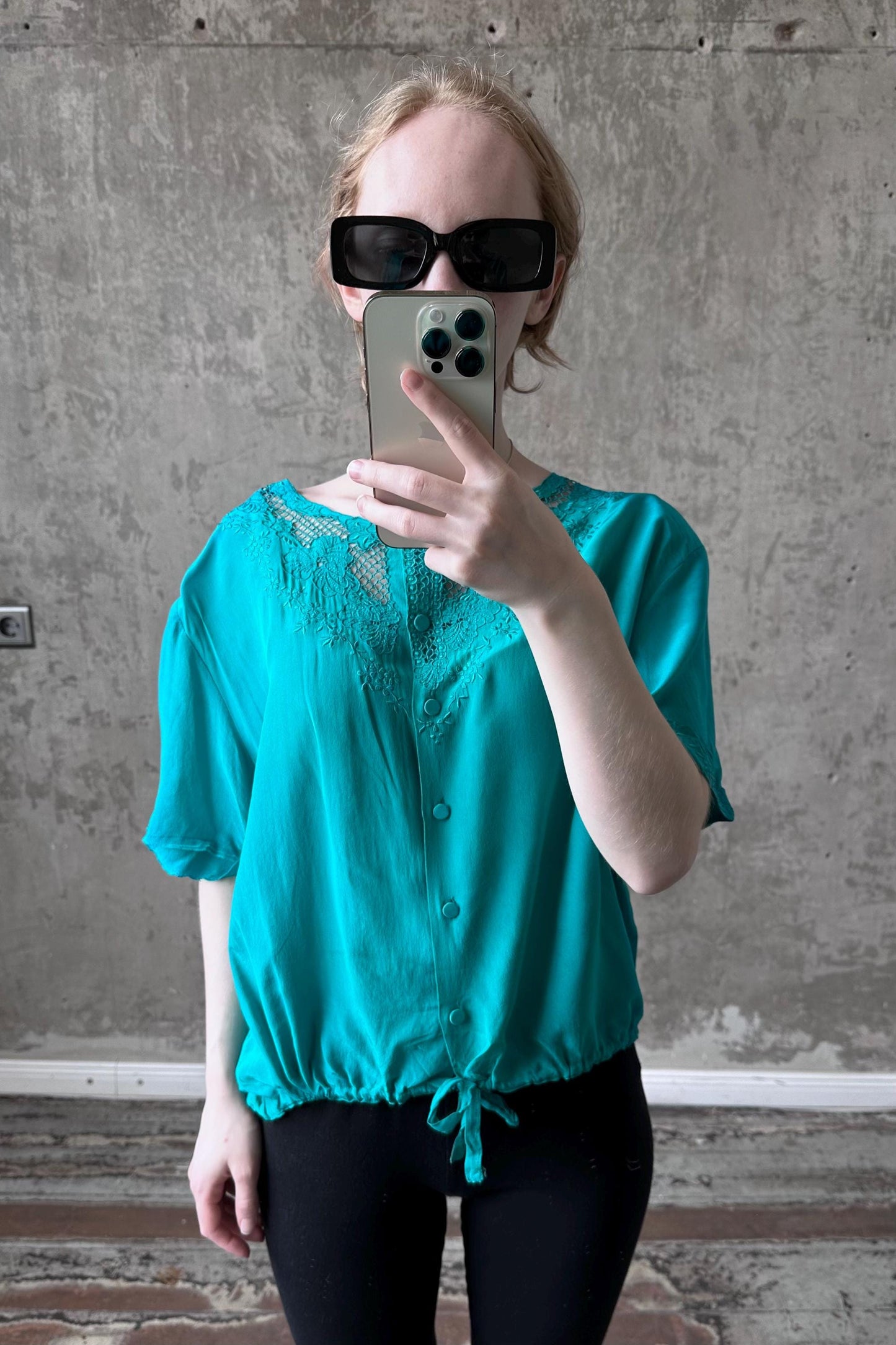 A woman wearing a turquoise blouse and sunglasses is taking a selfie with her iPhone.