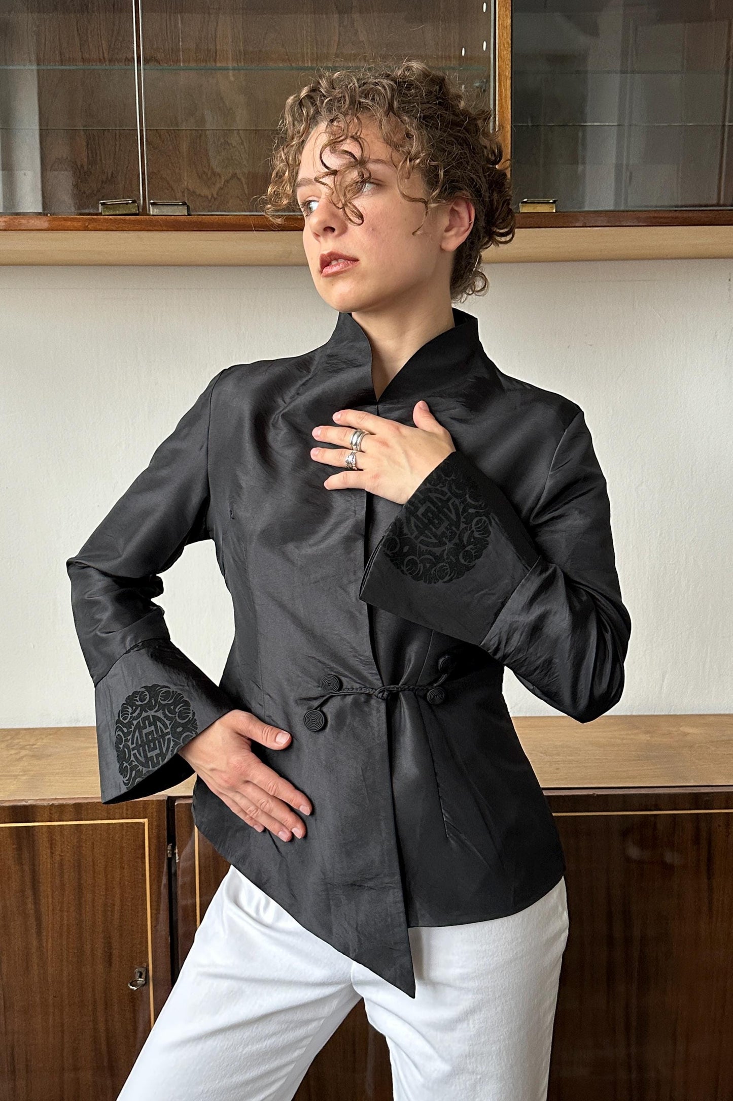 A woman with curly hair wearing a black jacket with a floral pattern stands in front of a wooden cabinet.