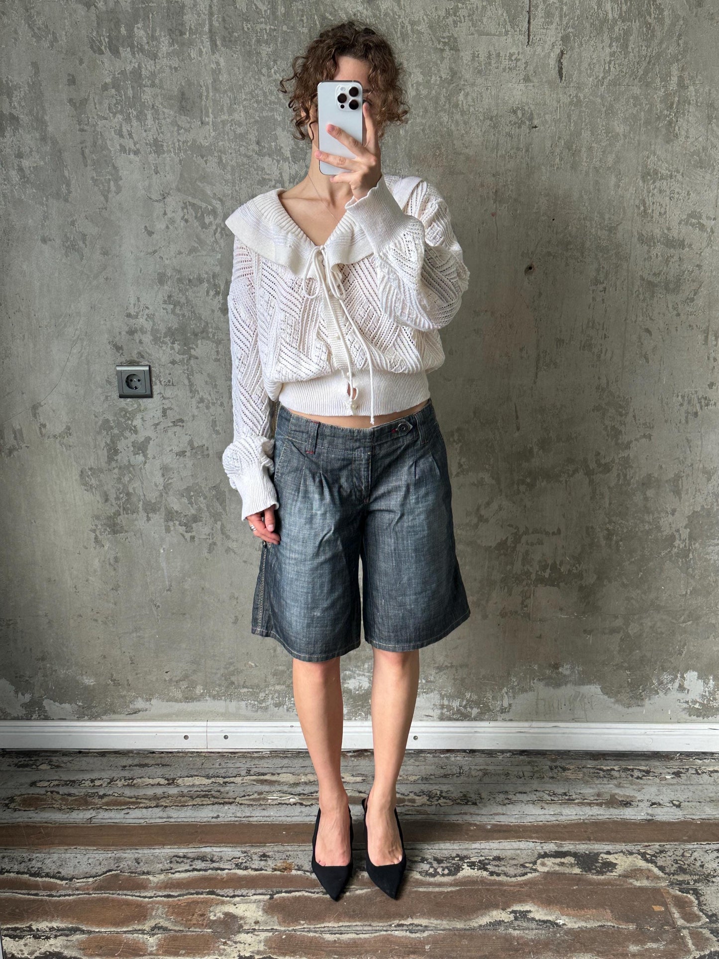 A person is taking a selfie with a white phone, standing in front of a concrete wall and wearing a white sweater, blue jeans, and black heels.